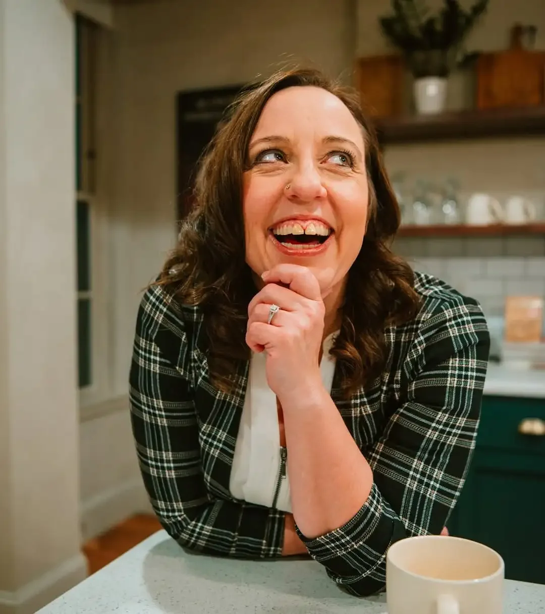 A woman with dark brown hair, smiling and looking up, sitting at a kitchen counter with a mug in front of her.