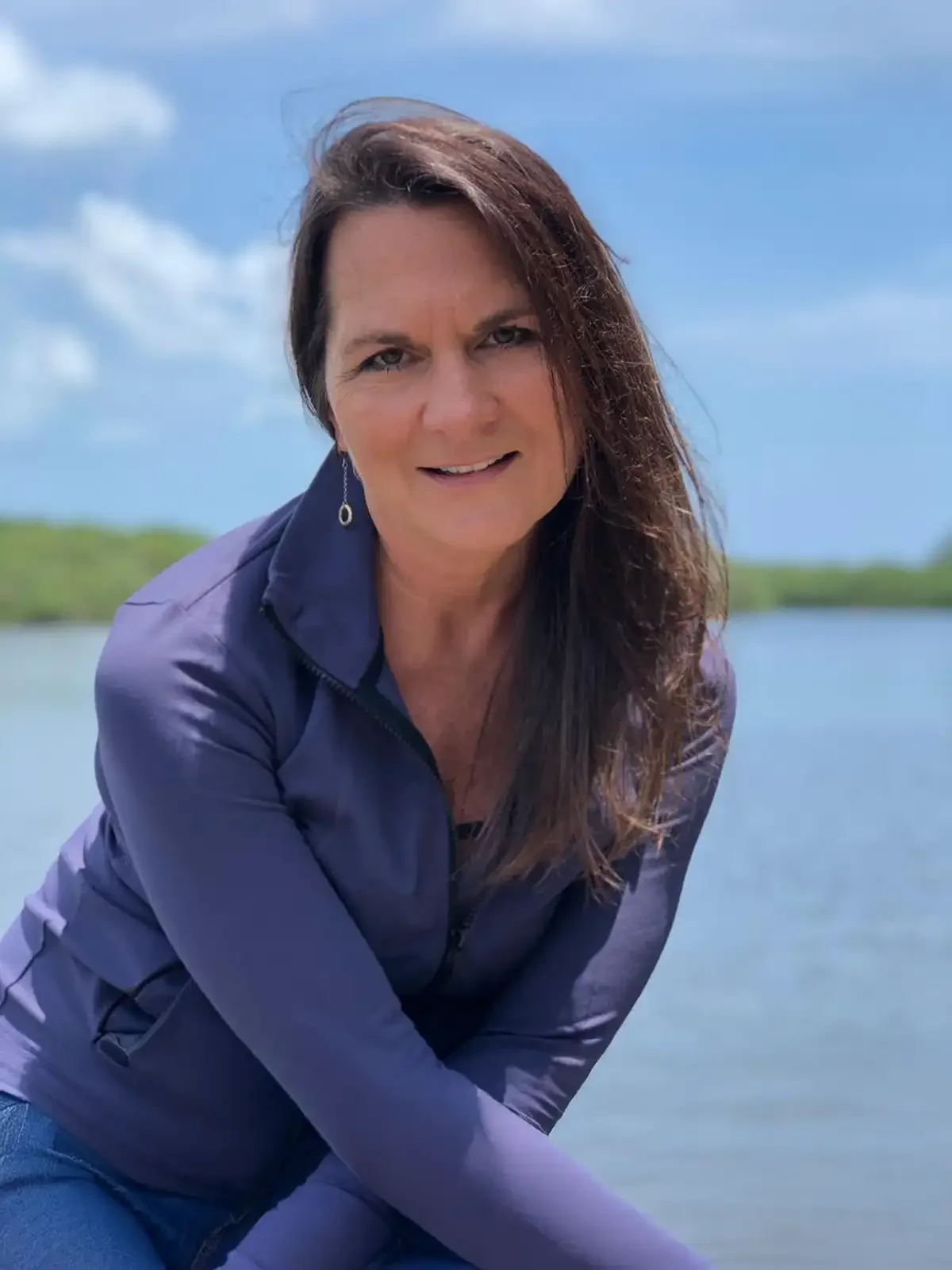 A woman with long brown hair wearing a purple jacket leaning forward outdoors by a body of water under a blue sky with clouds.