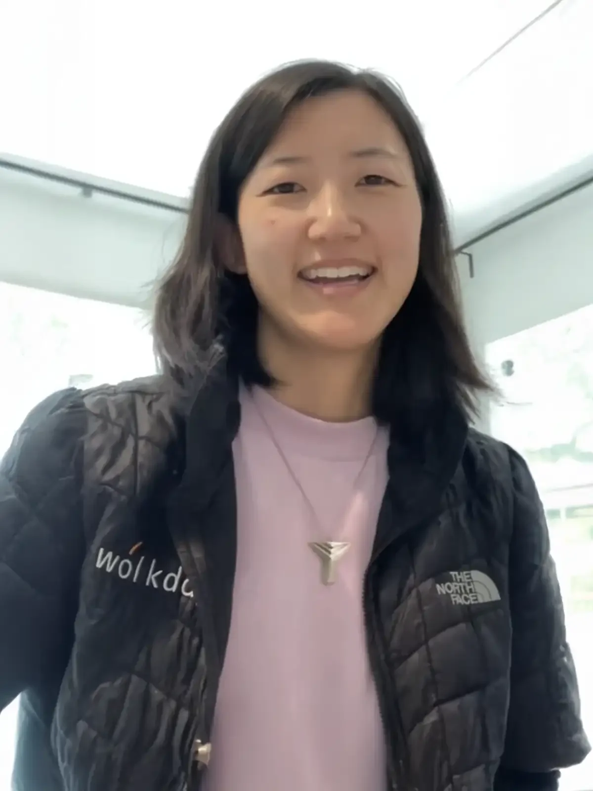A woman with dark hair smiling, wearing a black North Face jacket over a pink shirt, and a silver necklace, standing indoors with large windows behind her.