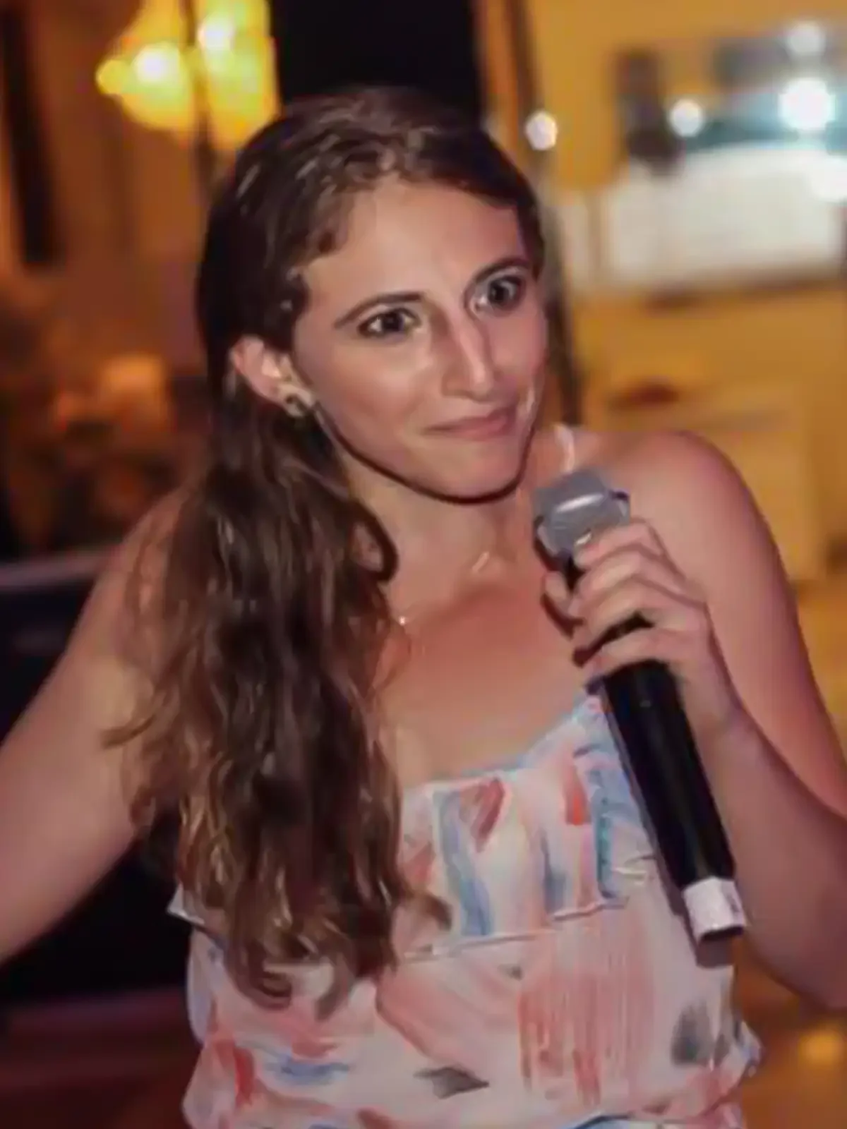A woman with long curly hair holding a microphone, appearing to be singing or speaking in a lively environment.