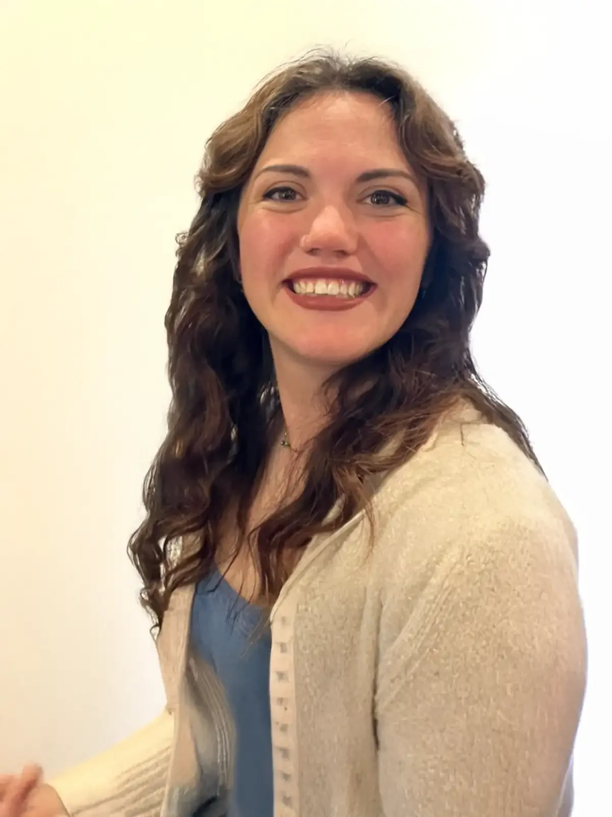A woman with long, curly brown hair smiling and showing her teeth, wearing a blue top and a beige cardigan, standing against a plain white background.