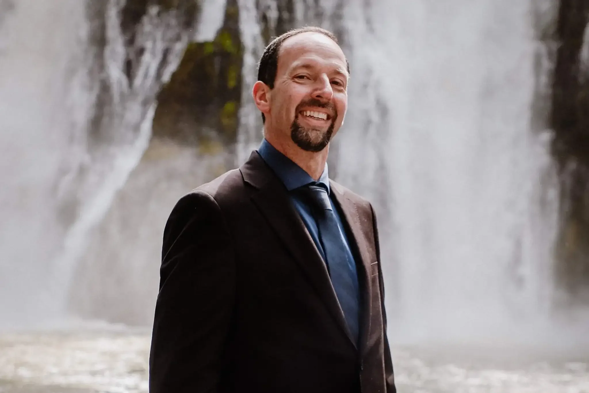 A man in business attire standing in front of a waterfall, smiling at the camera.