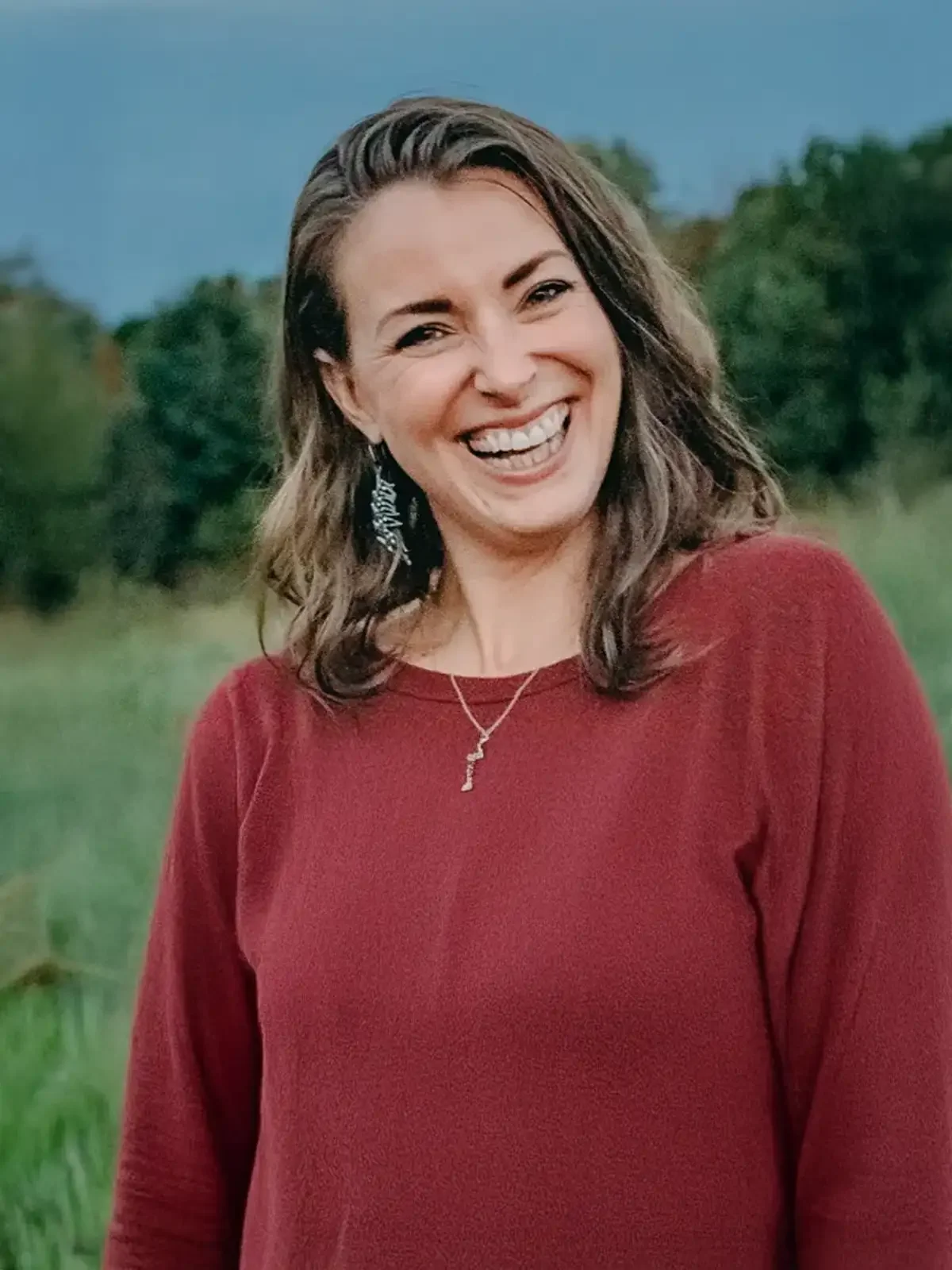 A woman with brown, wavy hair smiling outdoors, wearing a red long-sleeve top, earrings, and a necklace with a letter "K" pendant, with trees and a grassy landscape in the background.