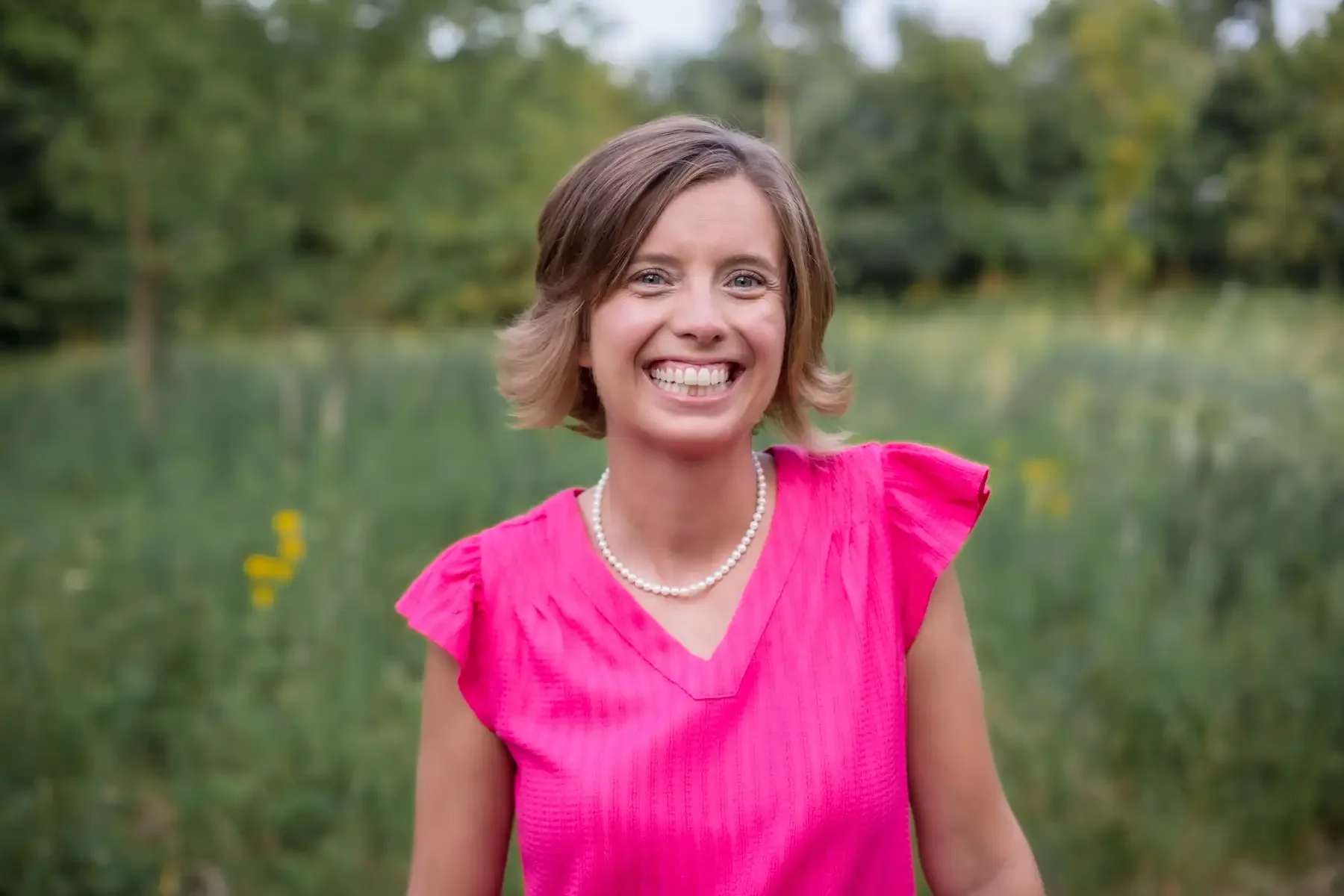 A woman with short light brown hair smiling outdoors in front of a pond surrounded by green trees, wearing a bright pink dress and a pearl necklace.