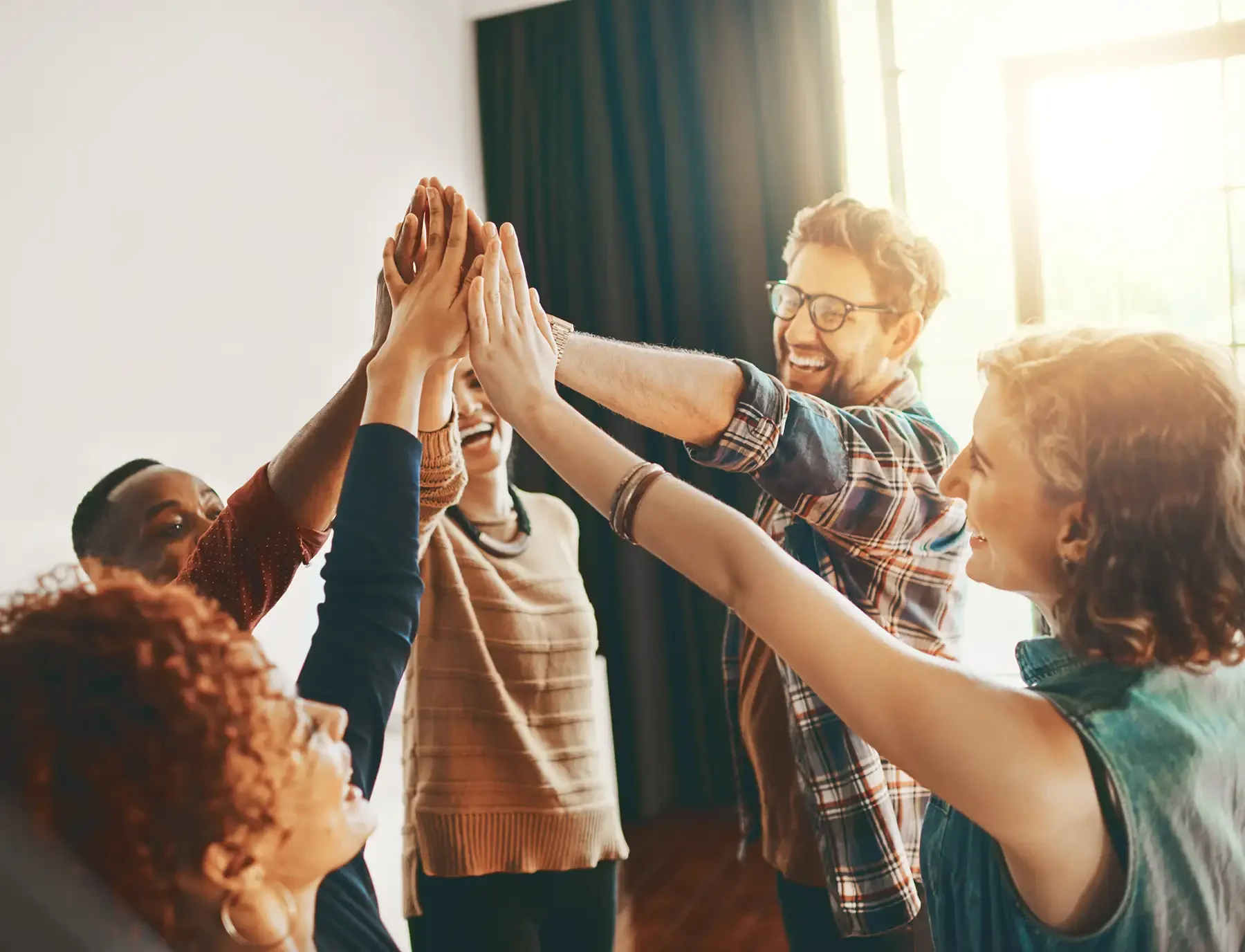 A group of diverse friends in a bright room celebrating with a high five, smiling and laughing.
