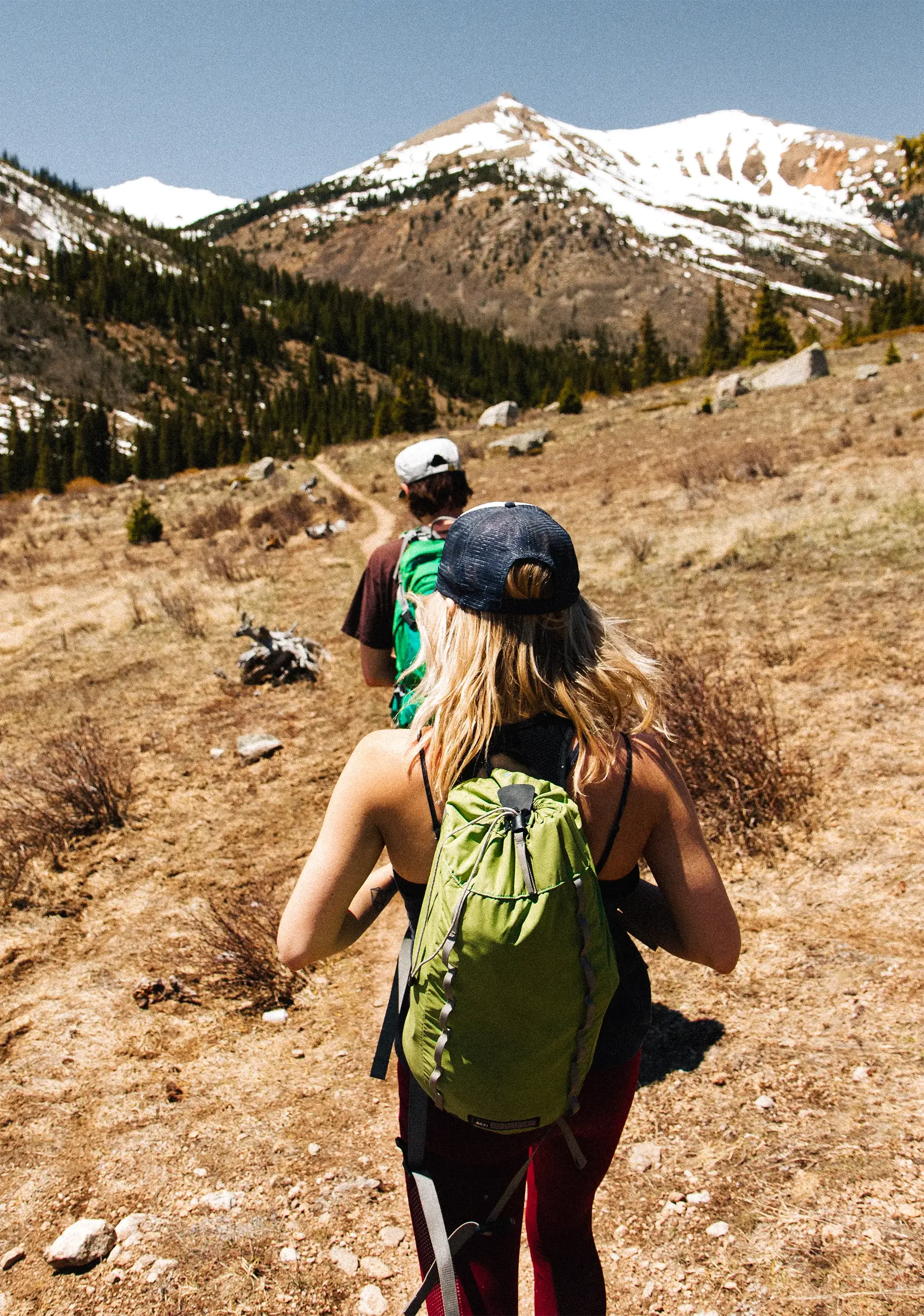 Two hikers with backpacks walking on a dirt trail through a mountainous area with snow-capped peaks and sparse vegetation.