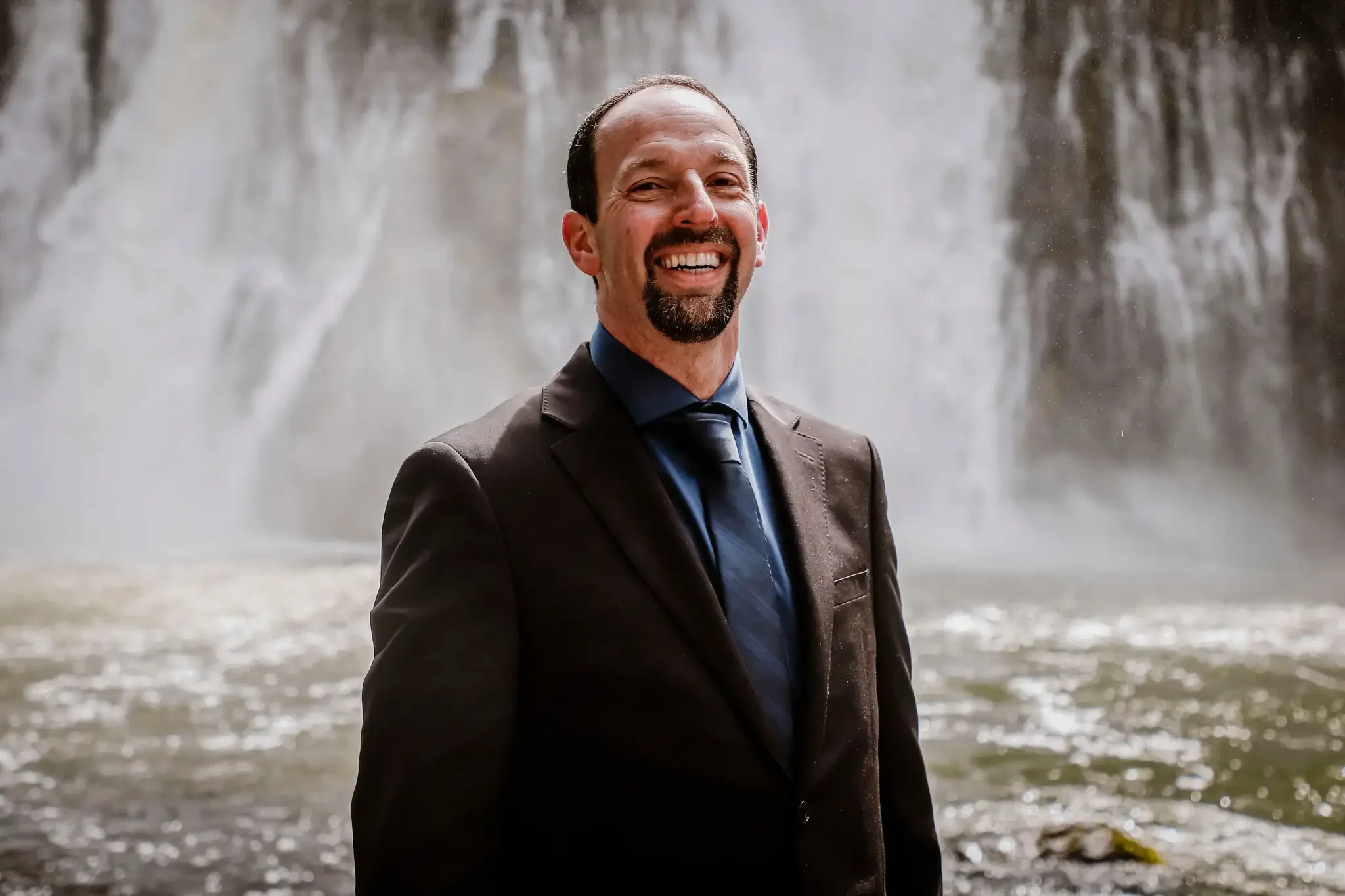 A man in a suit smiling outdoors near a waterfall.