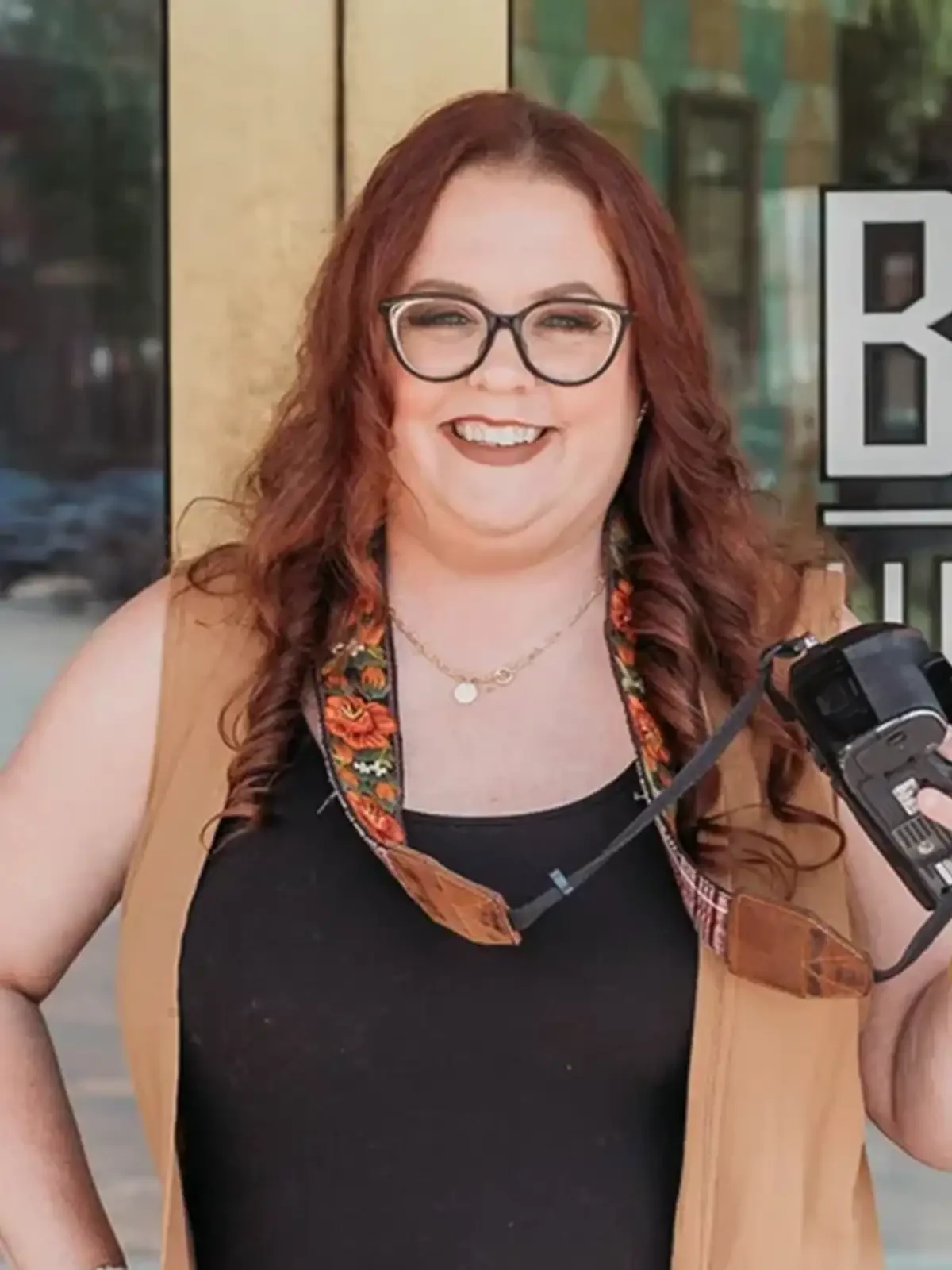 A woman with red hair, glasses, and a black shirt holding a camera outside a building with a partial sign.