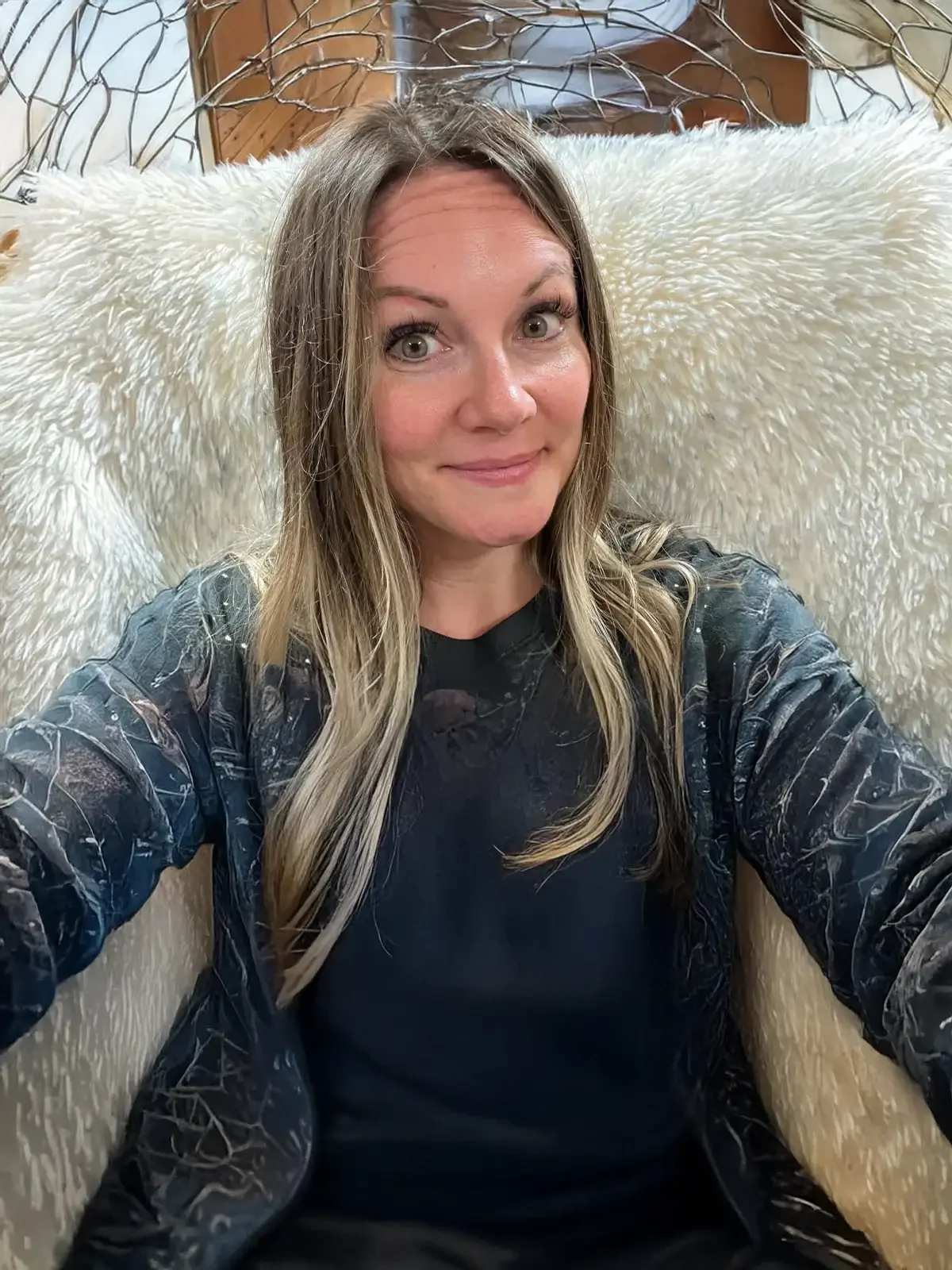 A woman with long blonde hair taking a selfie while sitting on a plush, cream-colored chair with a wireframe backrest, in a cozy room with wood paneling.