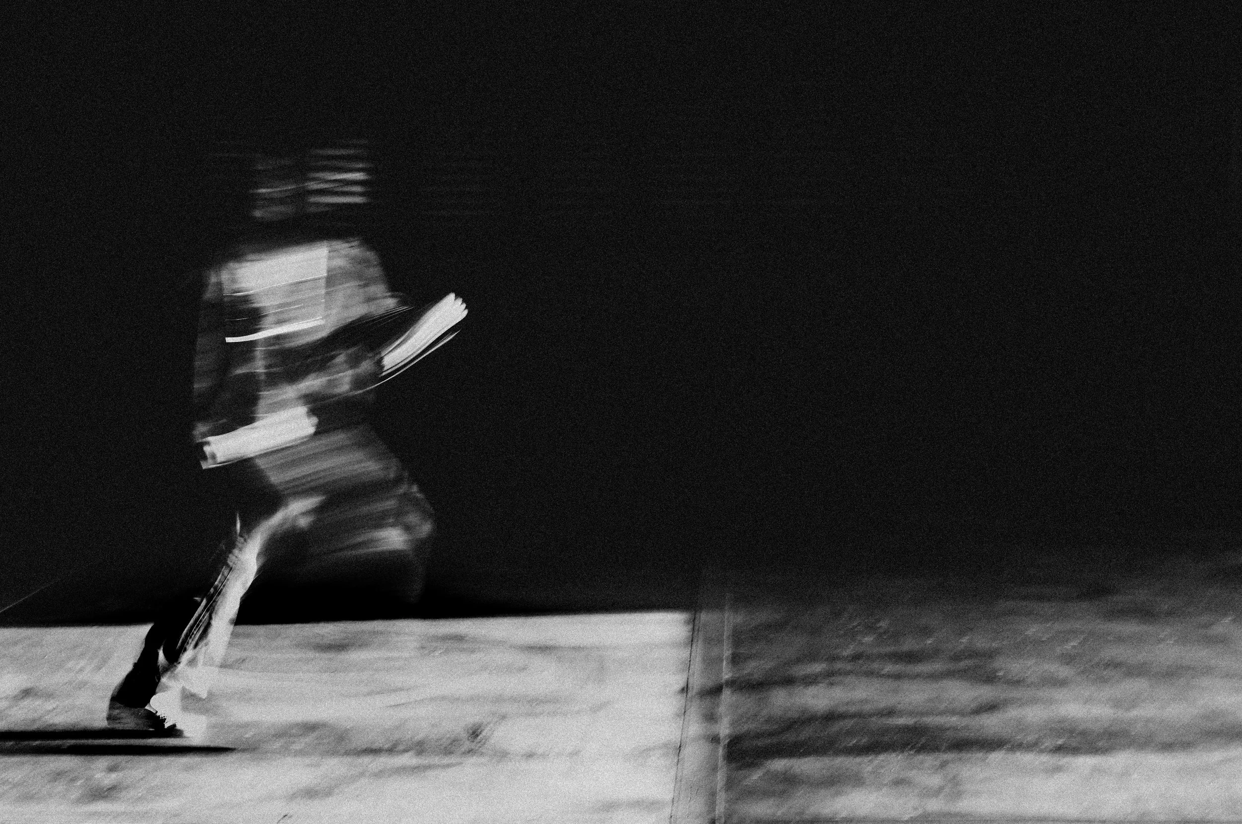 A black-and-white photo of a person running at night while holding a skateboard, with motion blur capturing the movement.