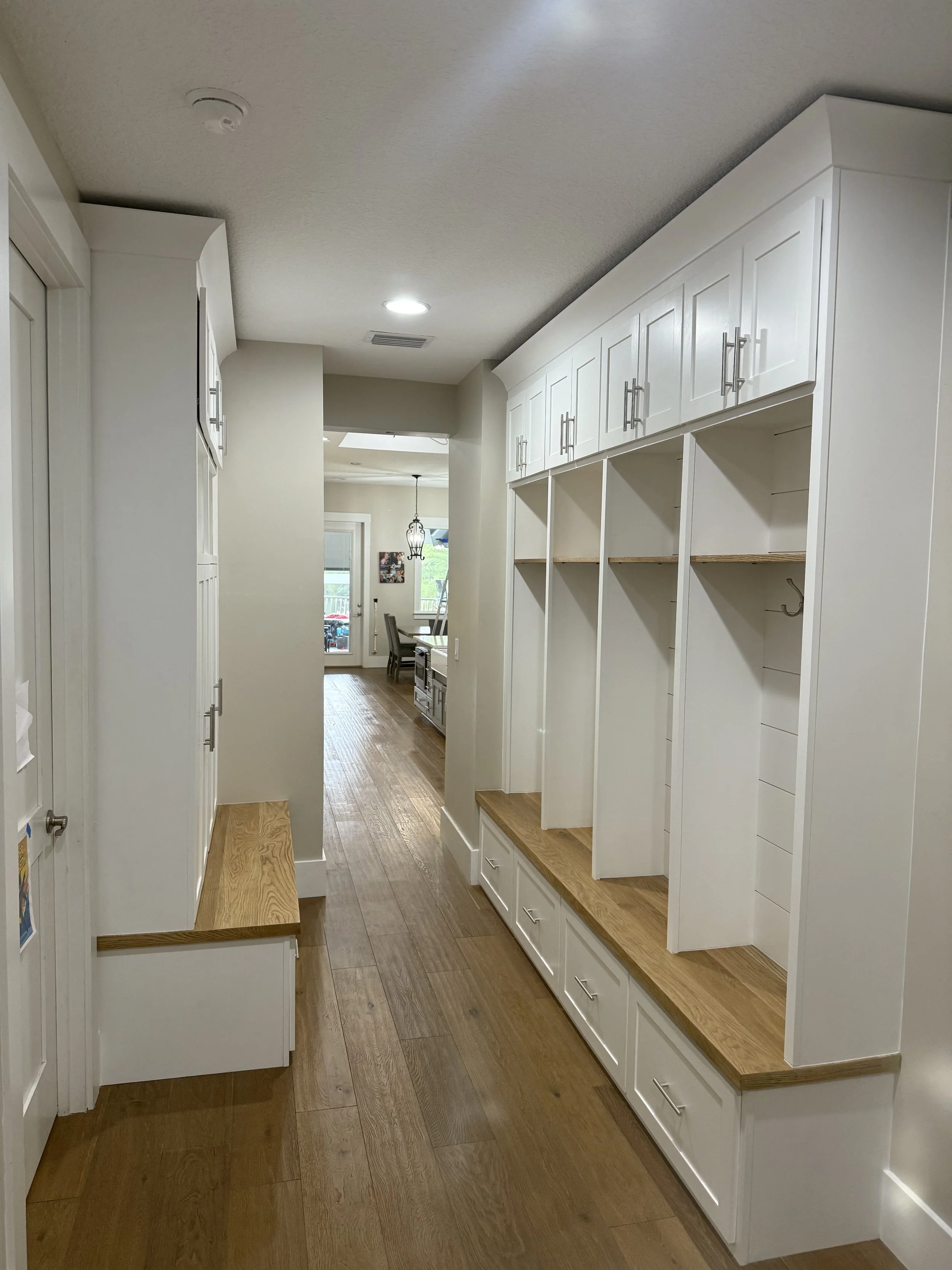 Interior view of a hallway with white built-in cabinets and drawers, hardwood flooring, leading to a dining area with a chandelier and a window.