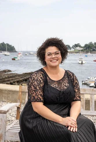 Woman with curly hair and glasses sitting on a bench by a body of water with small boats, wearing a black lace top and smiling.