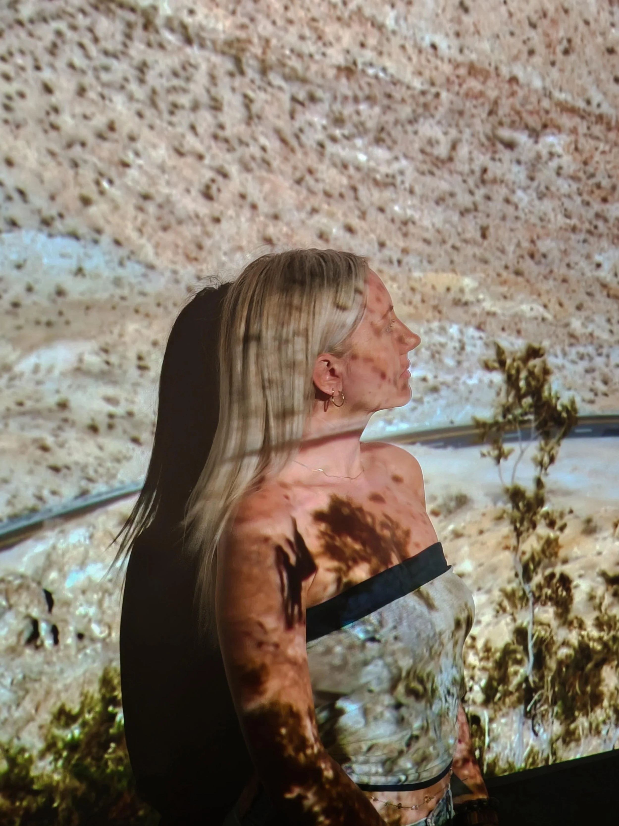 A woman with blonde hair and a strapless top stands in front of a screen displaying a desert landscape, with her profile visible and shadows of trees cast on her face and body.
