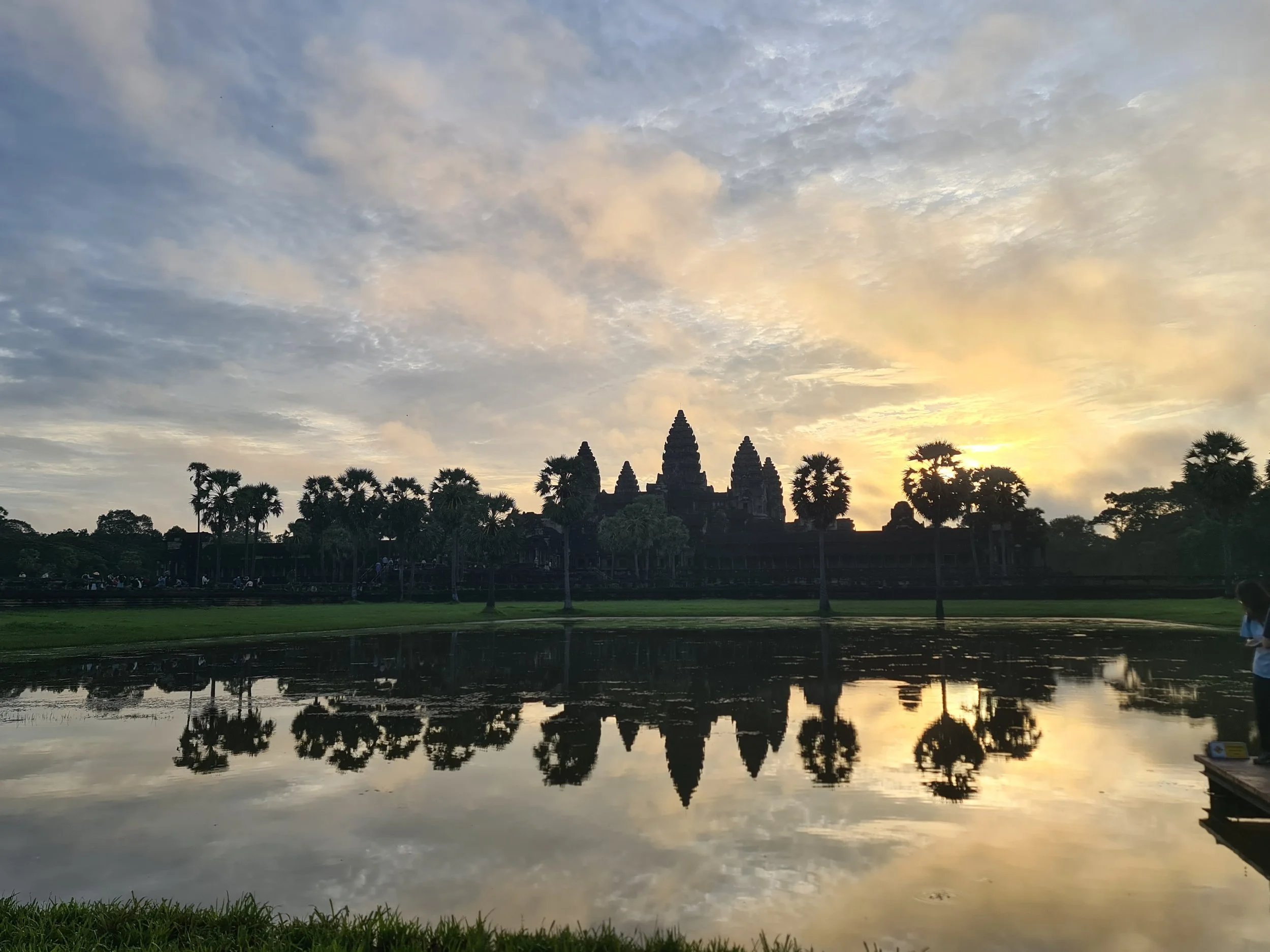 Sonnenuntergang hinter den Silhouetten der Tempelanlagen und Palmen, reflektiert im Wasser vor einem See.