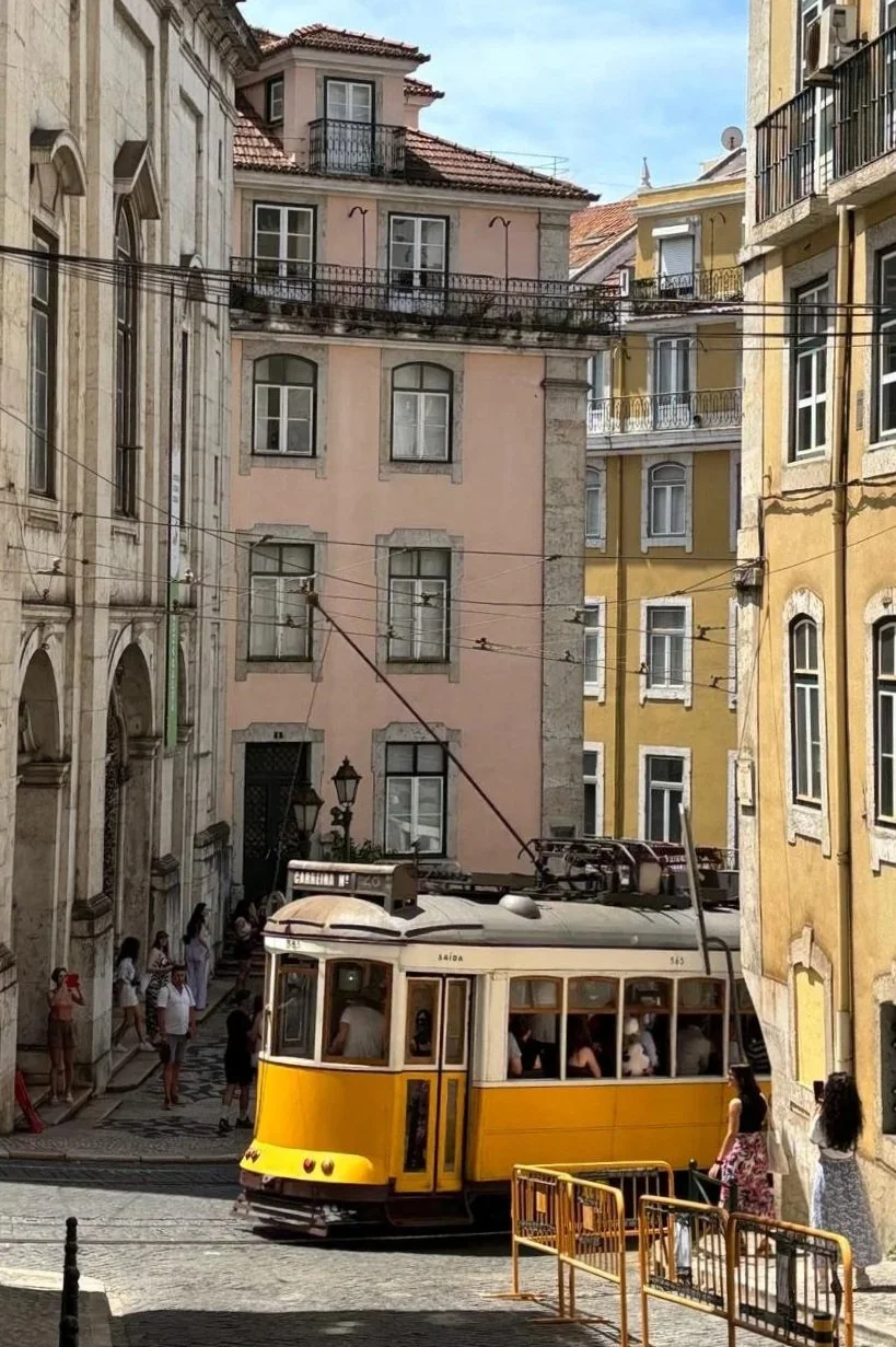 Ein gelber Straßenbahnwagen fährt durch eine enge, historische Stadtstraße, umgeben von bunten Gebäuden mit mehreren Etagen und Balkonen. Es gibt Fußgänger auf dem Bürgersteig, einige stehen und beobachten die Straßenbahn.