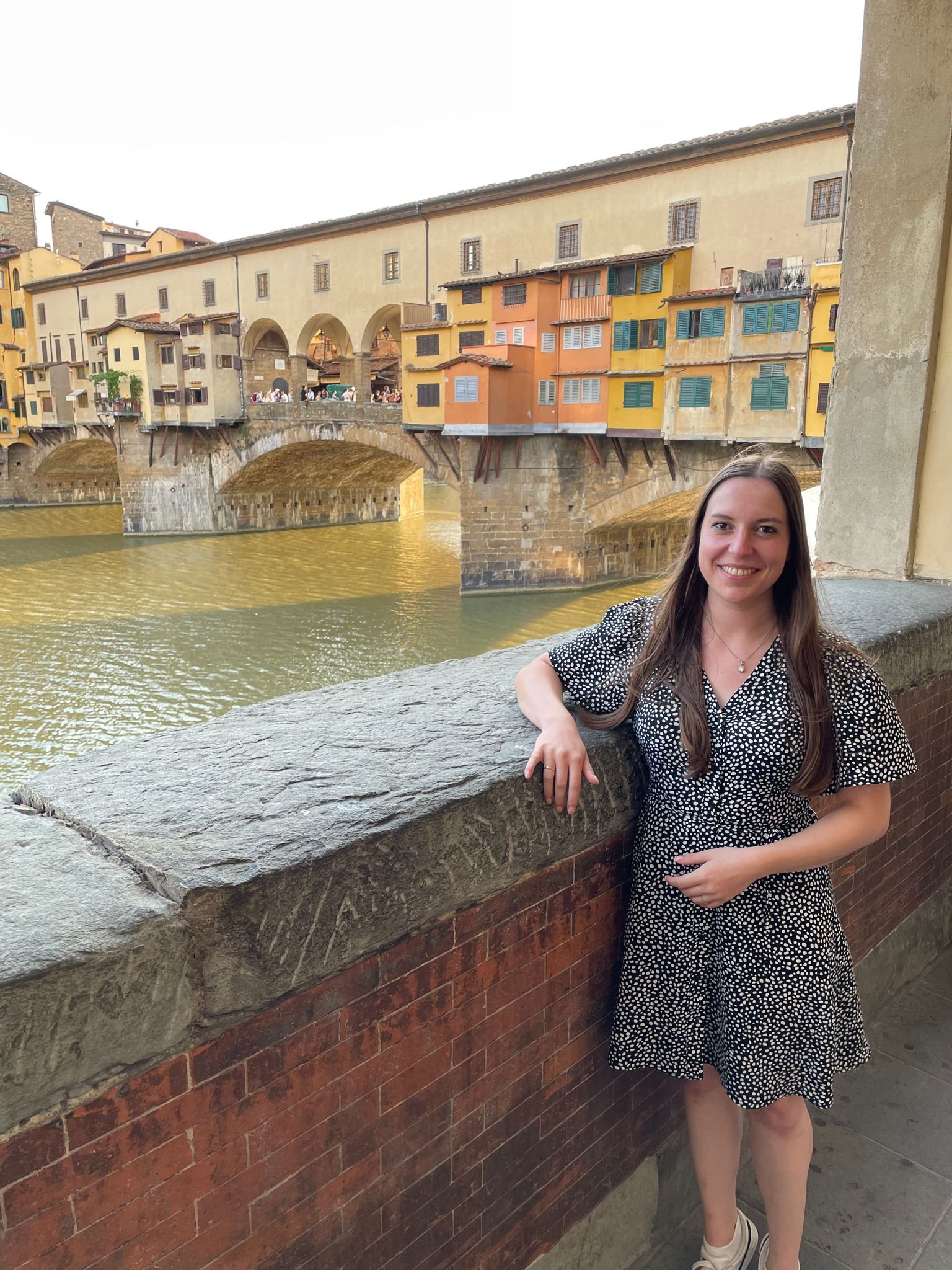 Eine lachende junge Frau steht an einer Steinmauer vor der Ponte Vecchio in Florenz, Italien, mit bunten Häusern und der Arno im Hintergrund.