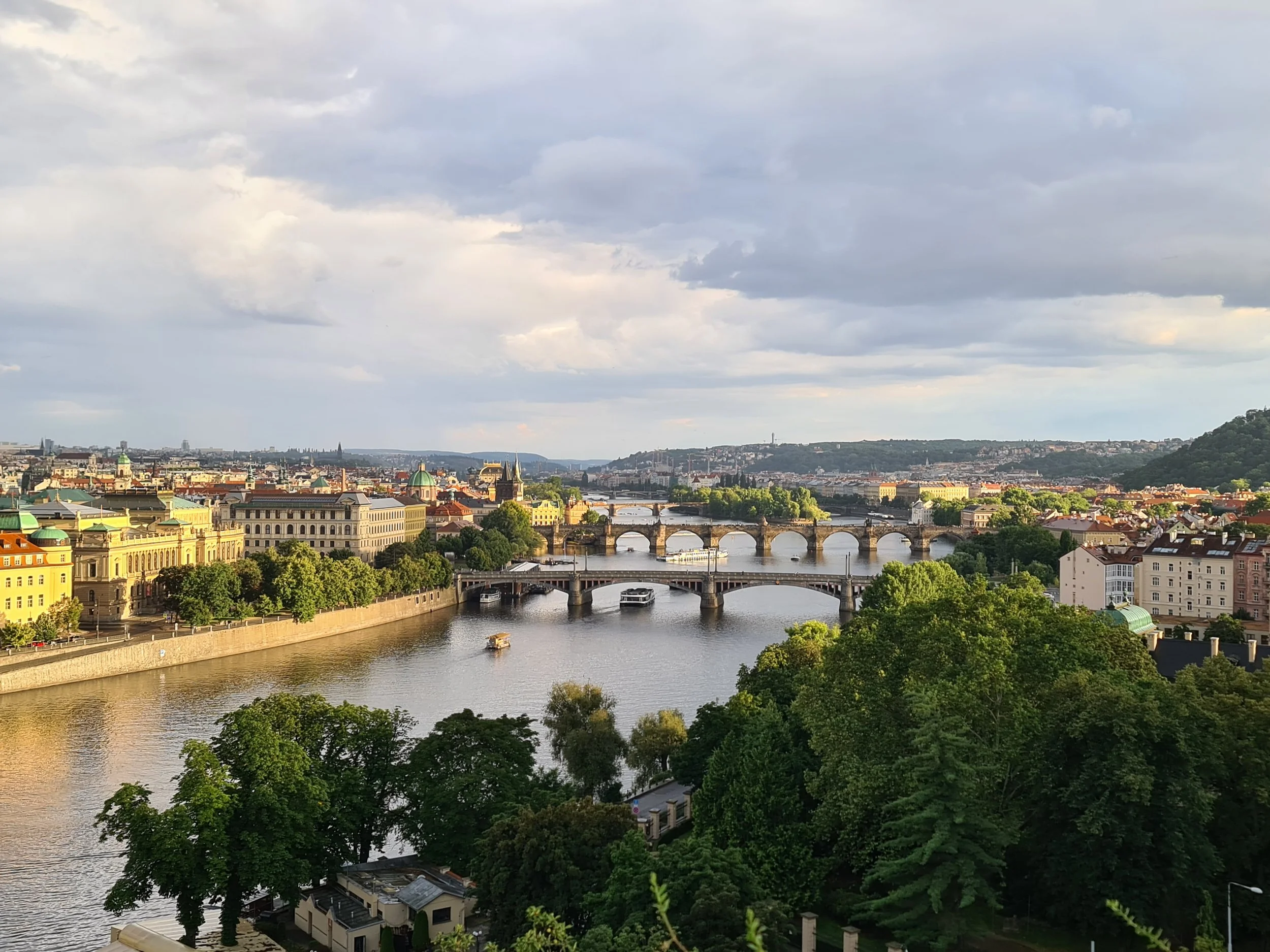 Landschaft mit Fluss, Brücken und Stadt im Sonnenlicht bei bewölktem Himmel.
