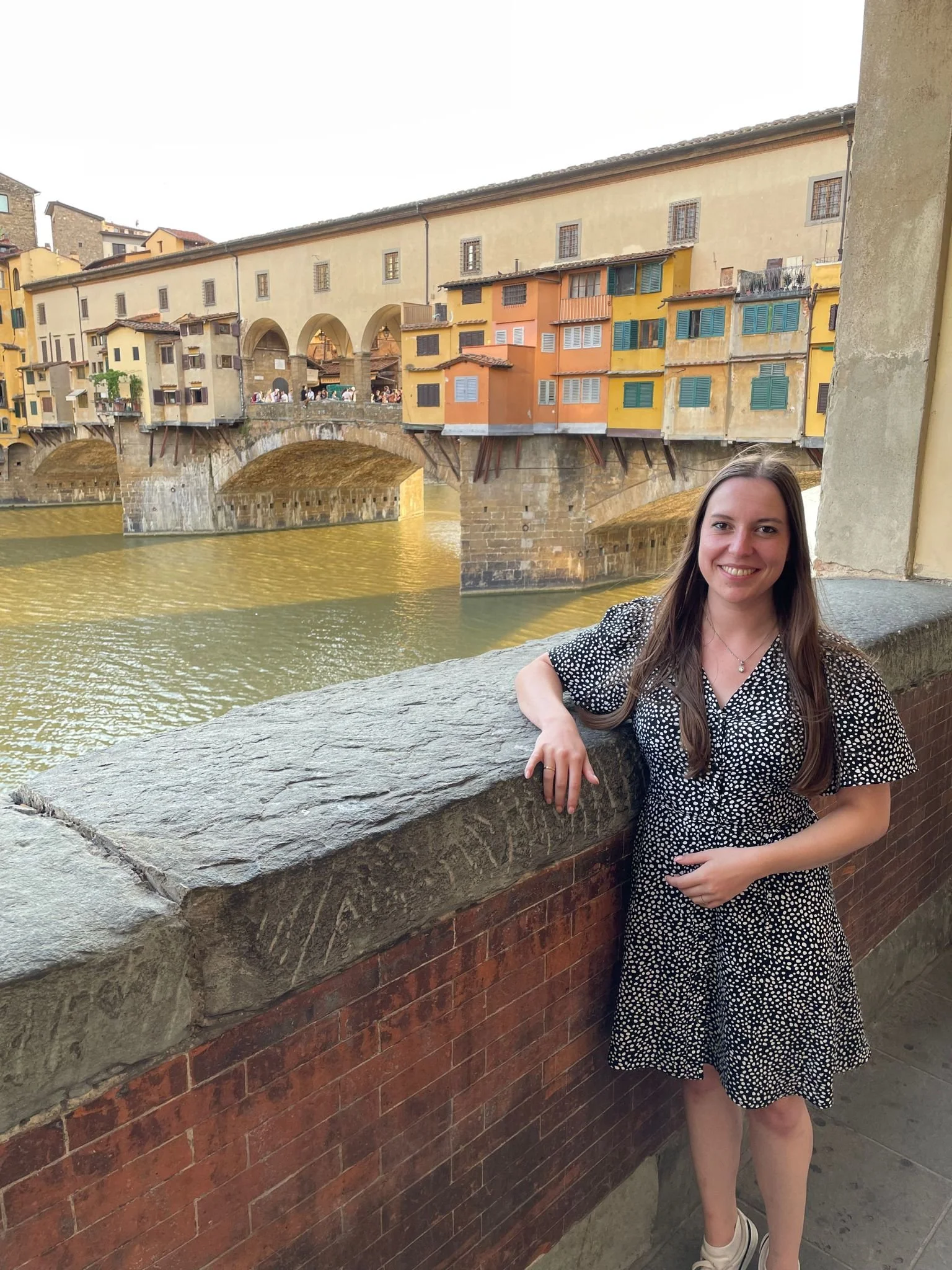 Eine junge Frau steht an einem Geländer mit Blick auf die Ponte Vecchio in Florenz, Italien, während sie lächelt.