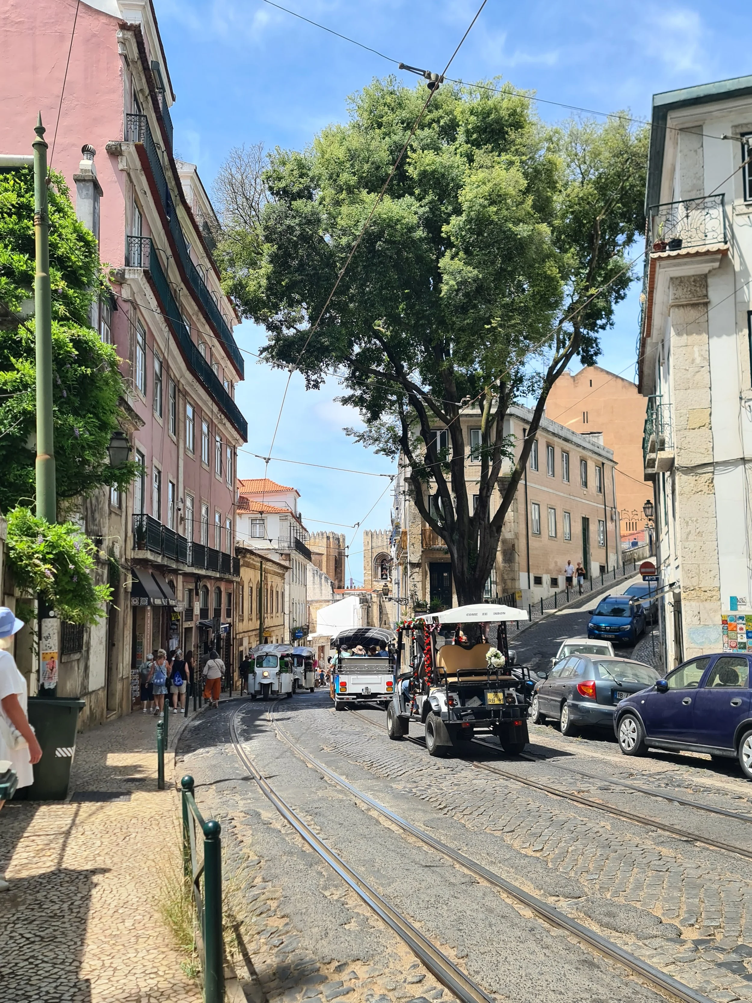 Straße mit Tramgleisen, einige Autos, u.a. ein Golf-Cart, und Fußgänger in einer malerischen, hügeligen Stadt mit alten Gebäuden, Bäumen und Kabeln am Himmel.