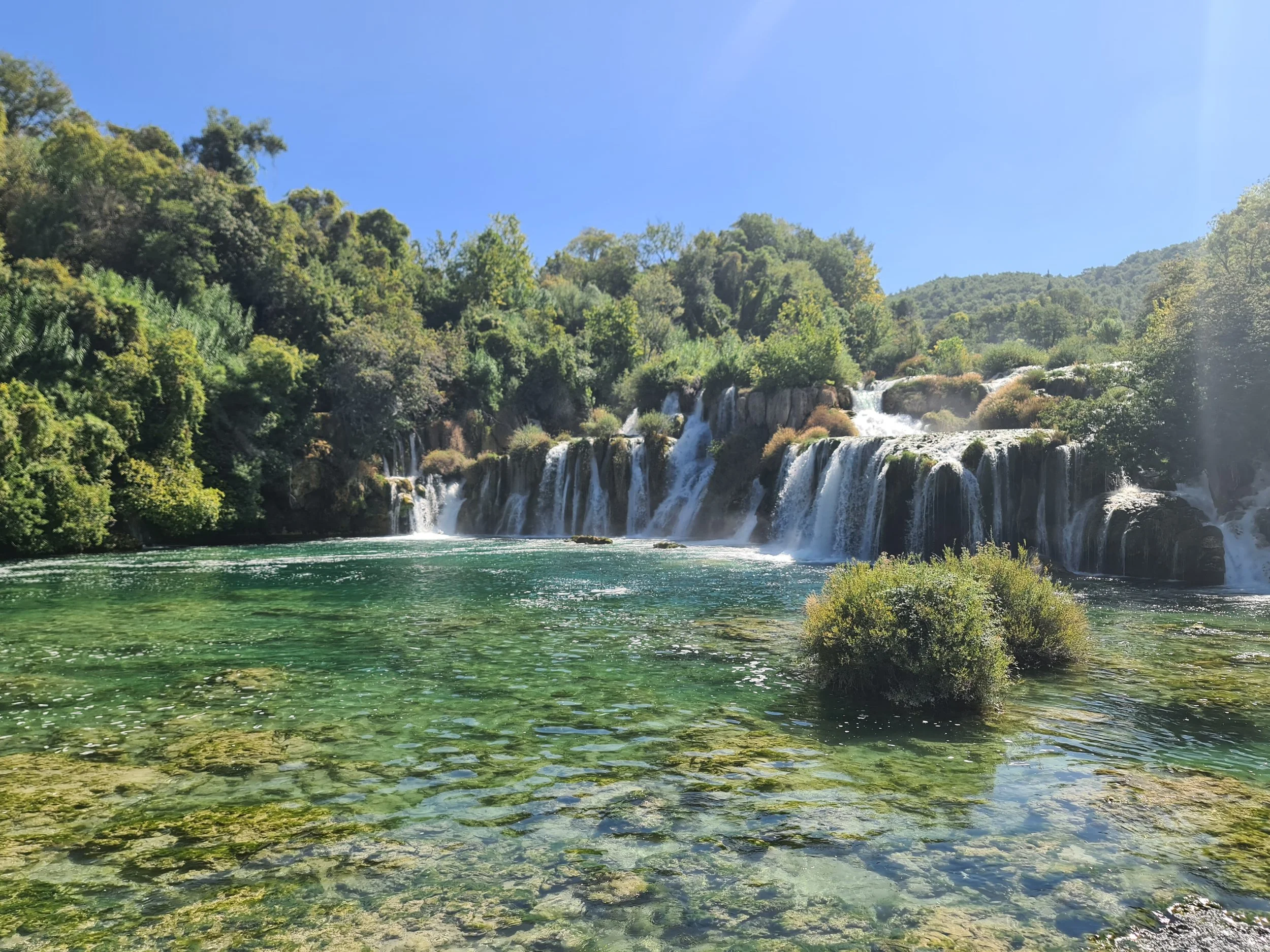 Blick auf einen Wasserfall mit mehreren kleinen Kaskaden, umgeben von üppigem grünen Wald und klarem, spiegelndem Wasser im Vordergrund. Himmel ist blau und sonnig.