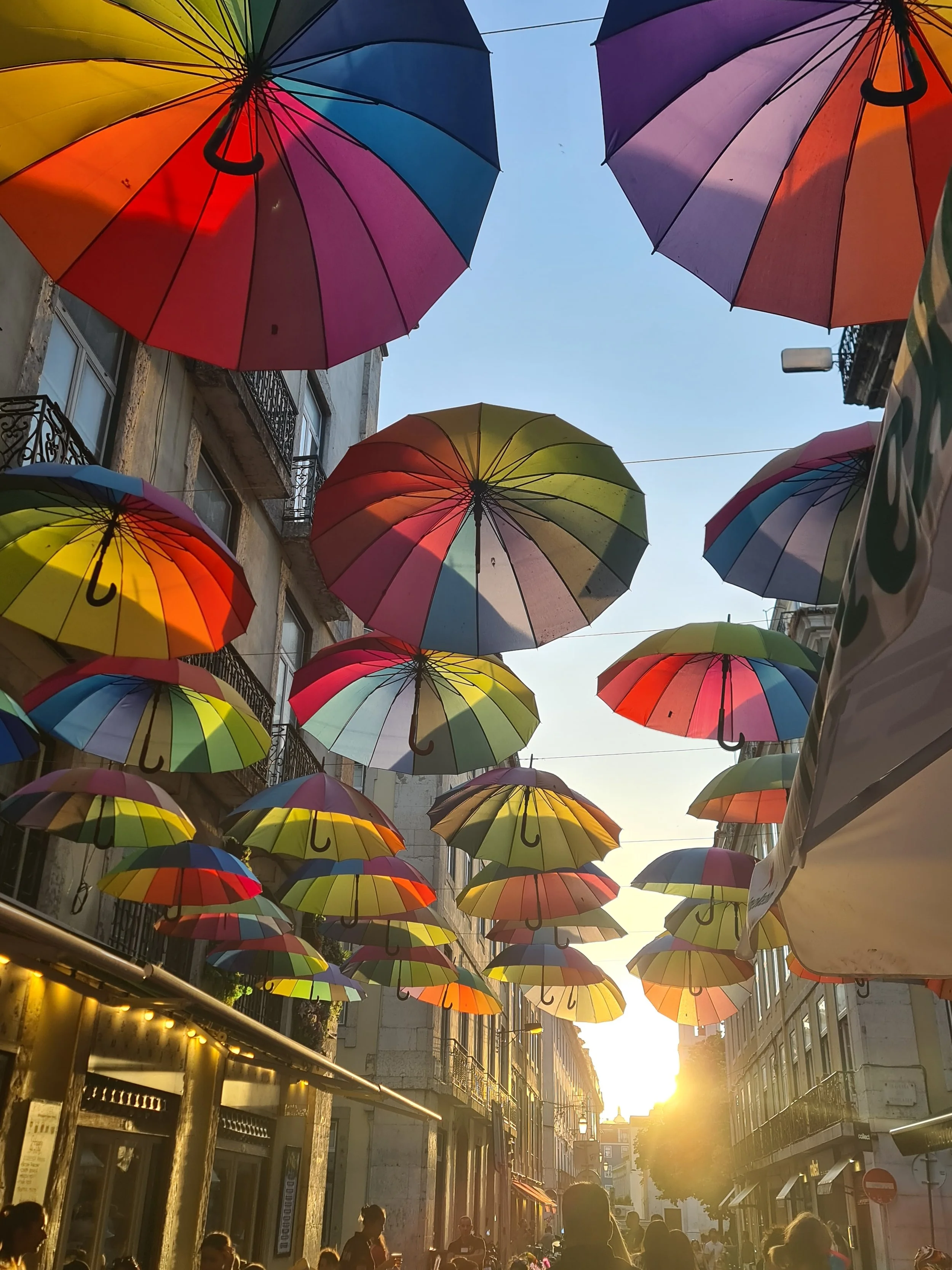 Viele bunte Regenschirme hängen über einer Straße bei Sonnenuntergang, während Menschen spazieren gehen.
