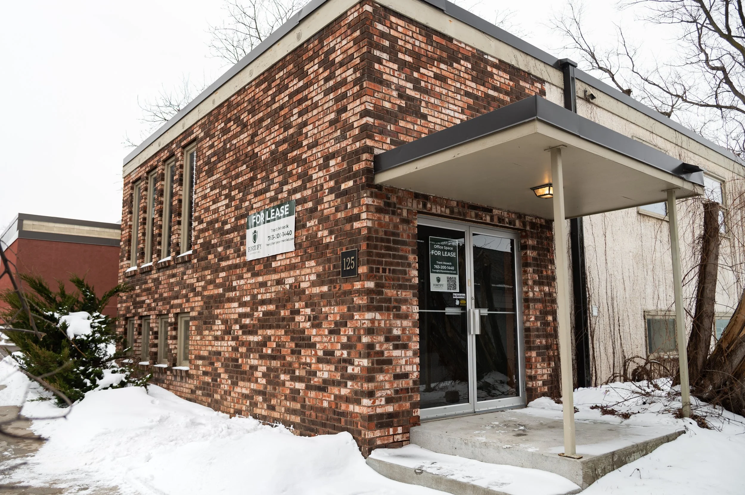 Empty shopping plaza with storefronts, glass doors, green awnings, and parking lot under a partly cloudy sky.
