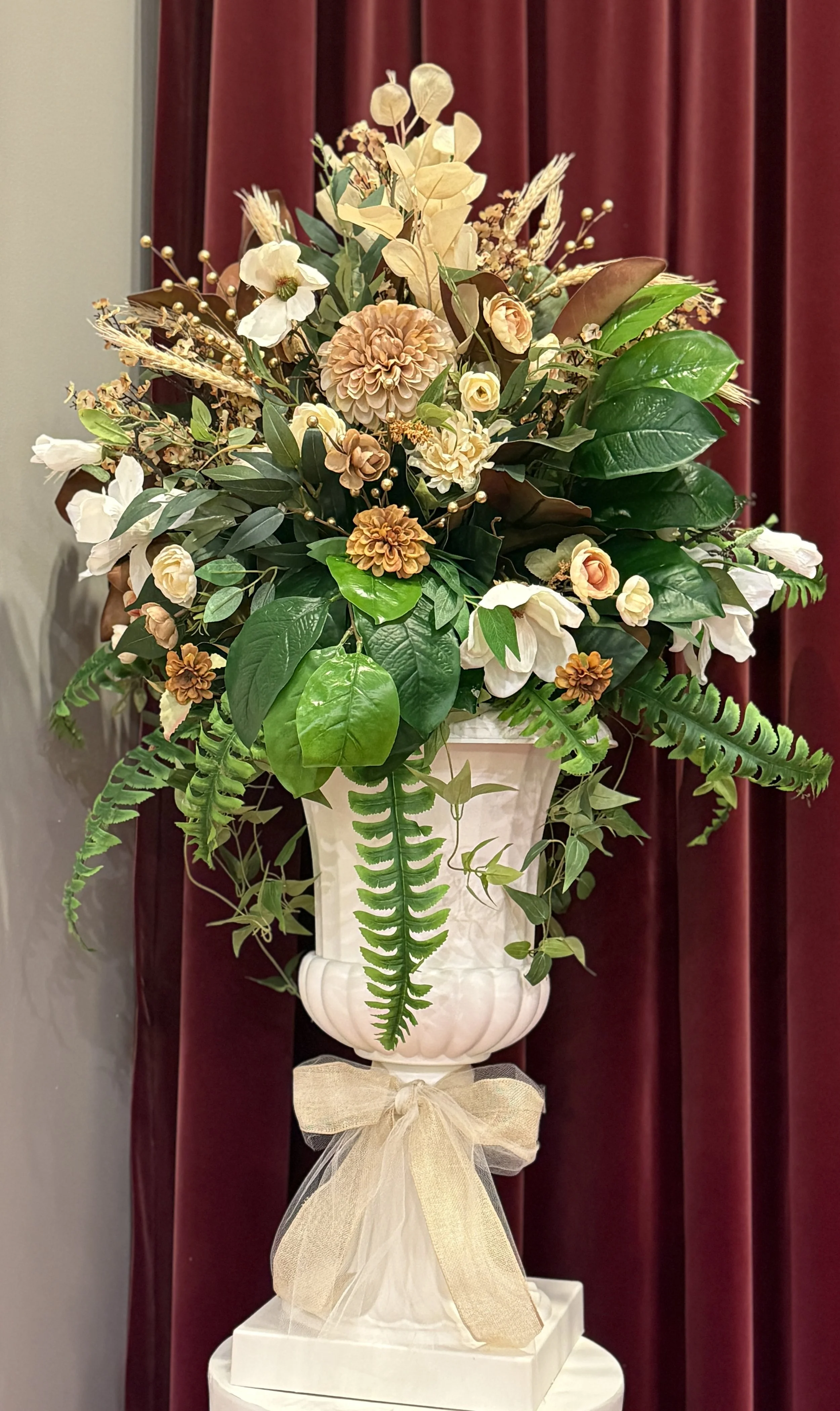 A floral arrangement with cream and brown flowers in a white urn decorated with a beige ribbon.