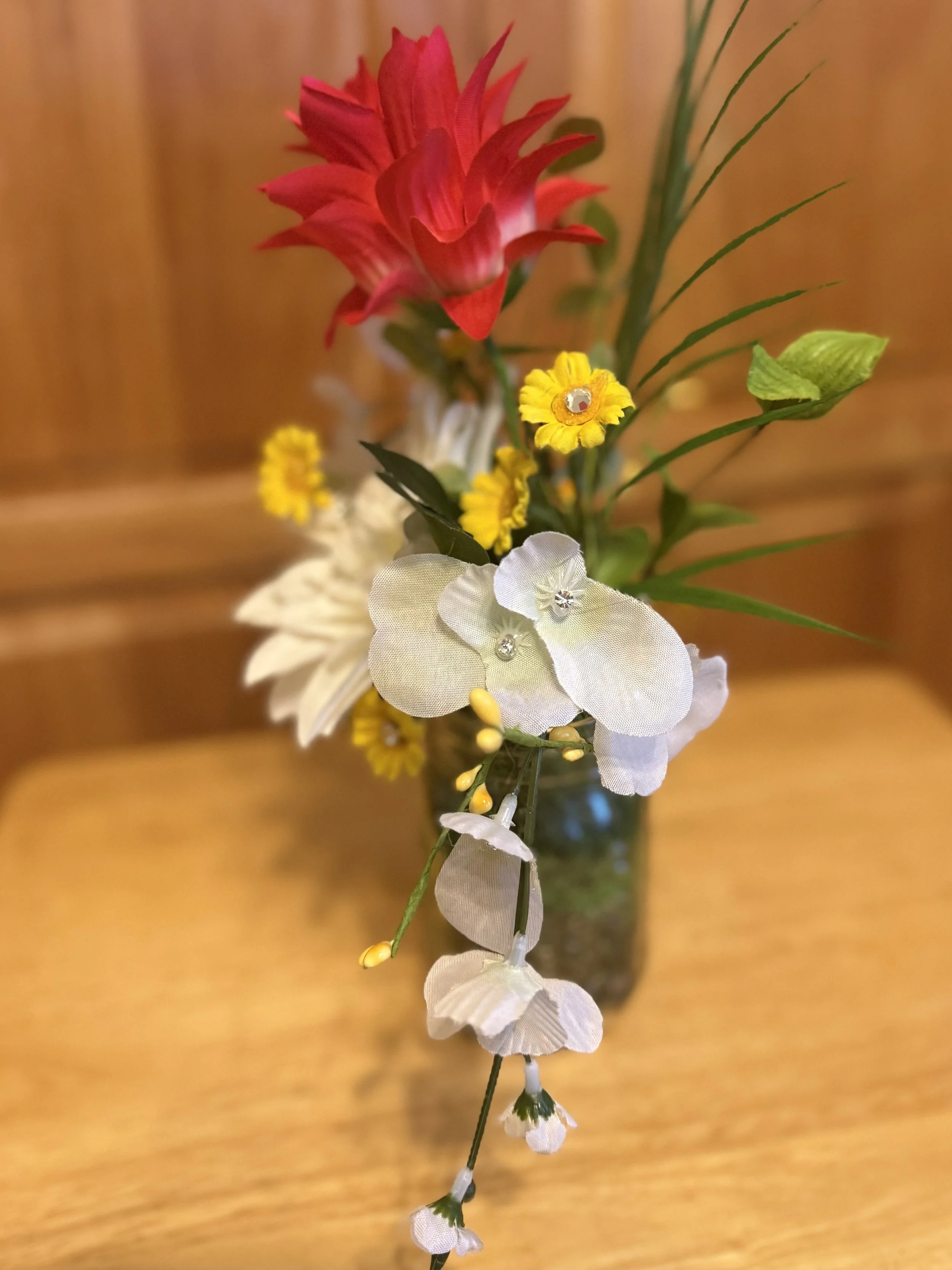 A floral arrangement in a glass vase containing a pink ginger flower, white orchids, yellow small flowers, white leaves, and green foliage, set on a wooden surface with a wood-paneled background.