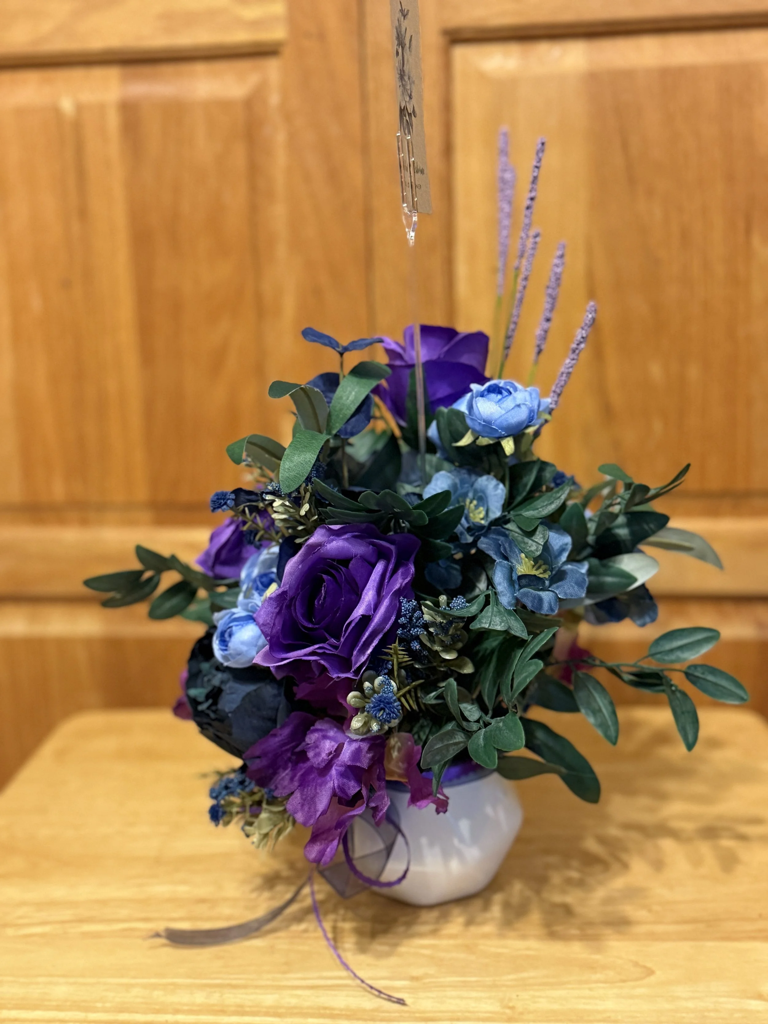 A small floral arrangement in a white vase, featuring purple roses, blue flowers, purple lavender, and green leaves, placed on a wooden surface against a wooden-paneled background.