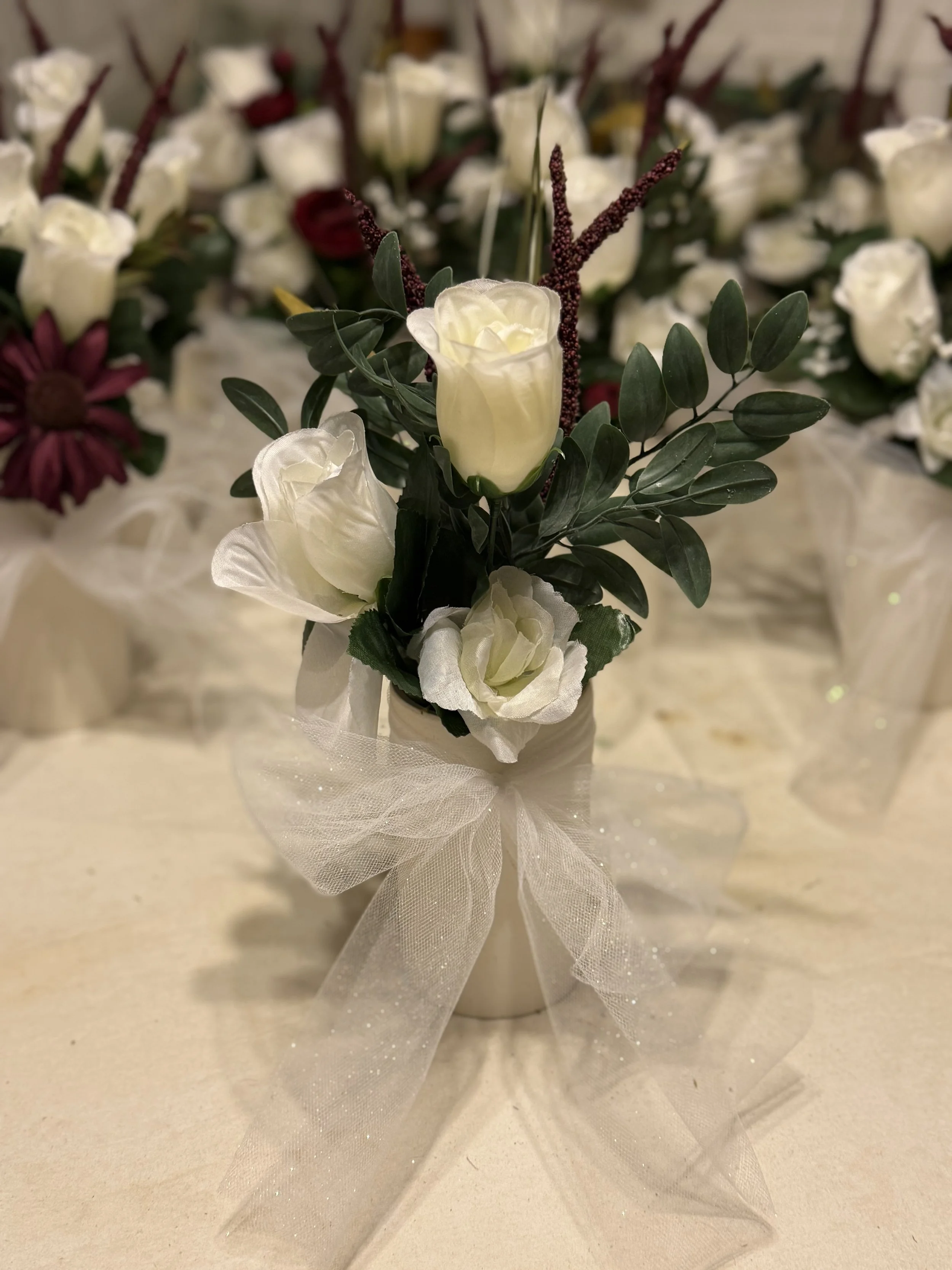A small floral arrangement with white roses, white flowers, green leaves, and burgundy accents in a white vase with a sheer ribbon bow, set on a light-colored surface.