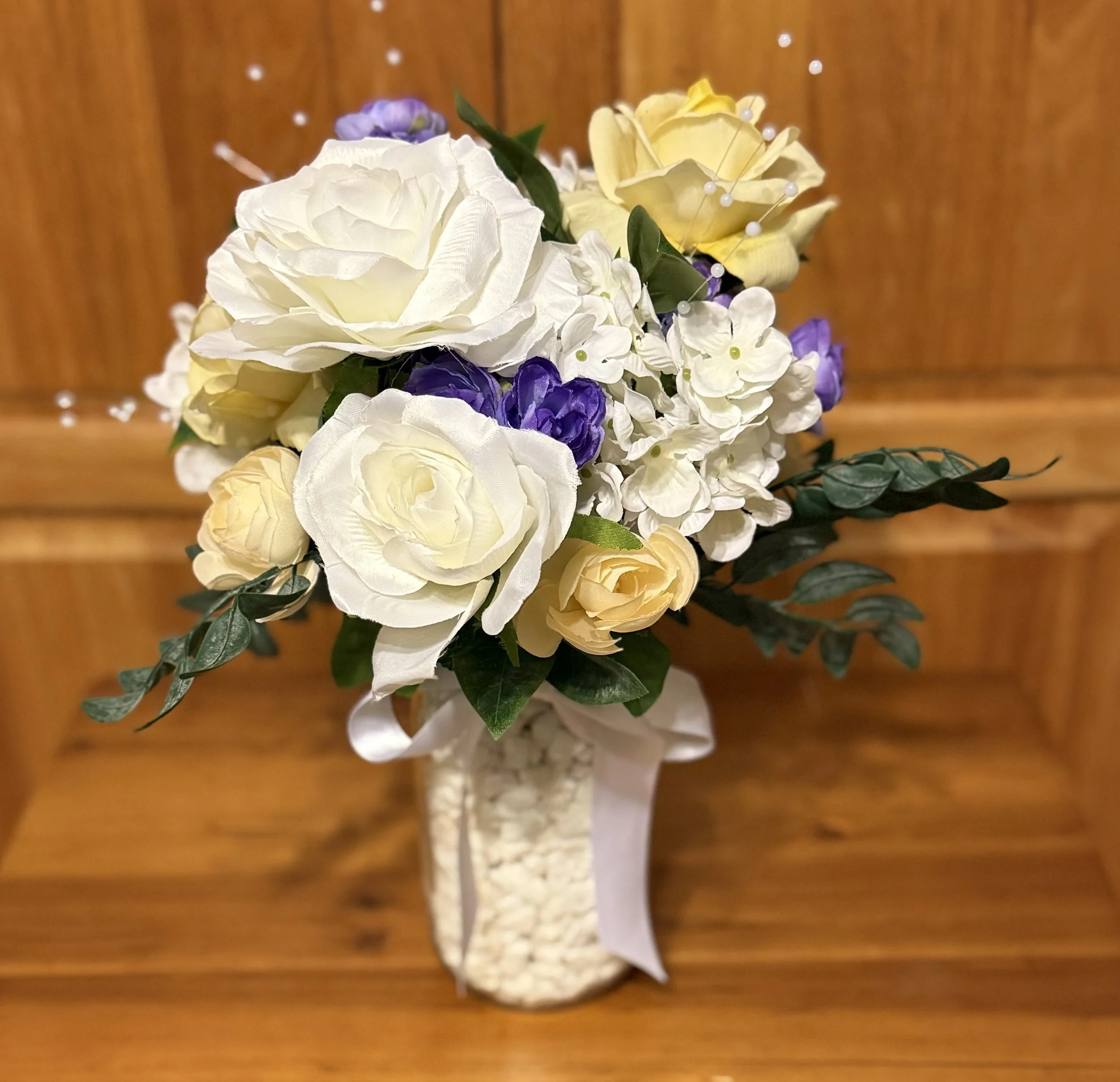 Artificial floral arrangement with white, yellow, and purple flowers in a white ceramic vase on a wooden surface.