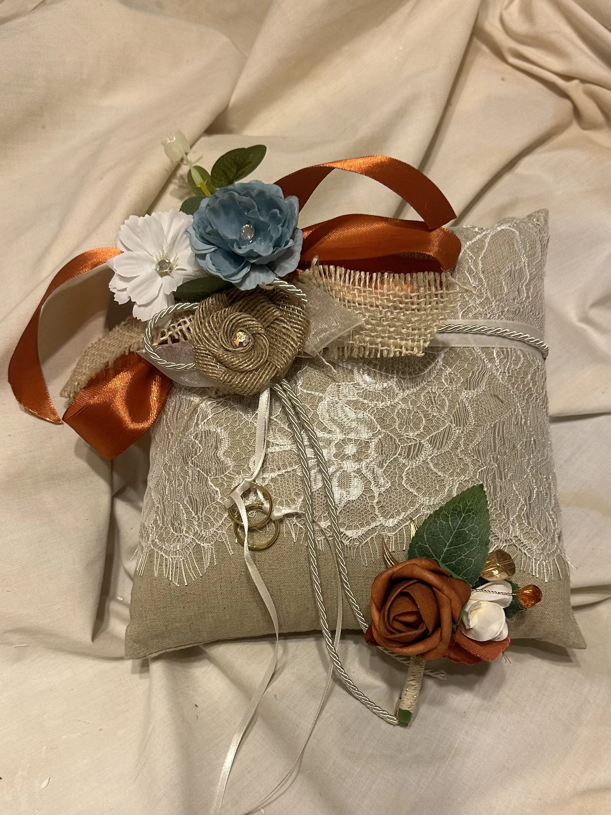 A decorative ring bearer pillow with lace and floral embellishments, including ribbons, fabric flowers, and rings, arranged on a light-colored fabric surface.