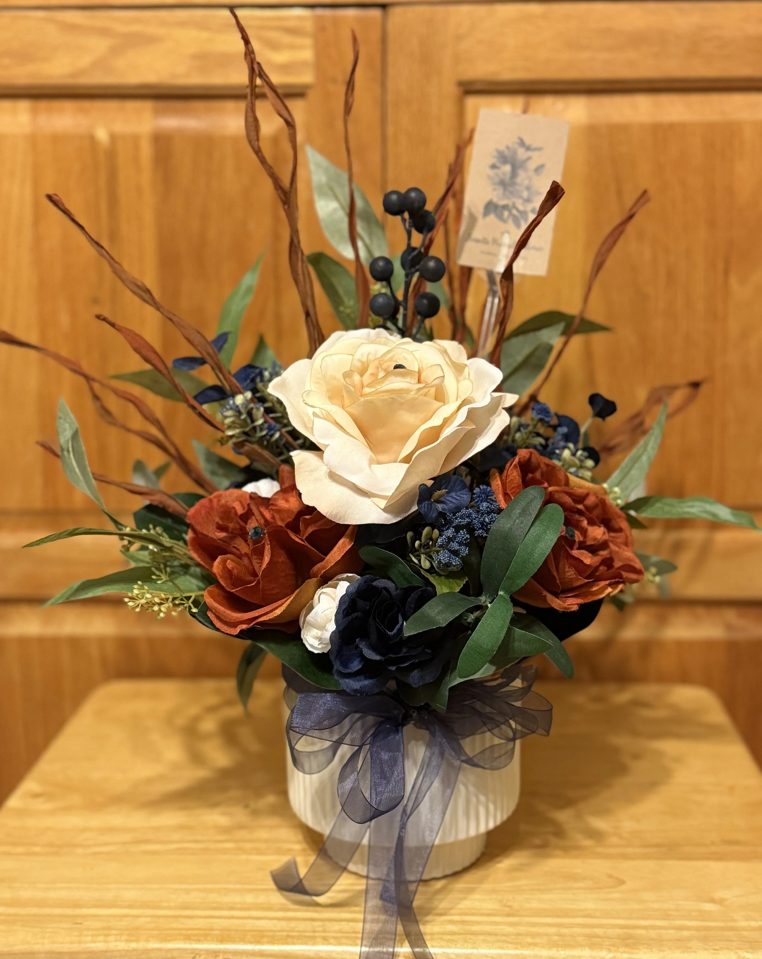 A floral arrangement with cream, rust, black, and blue flowers in a black vase, decorated with a black ribbon, surrounded by greenery and dried twigs, in front of a wooden background.