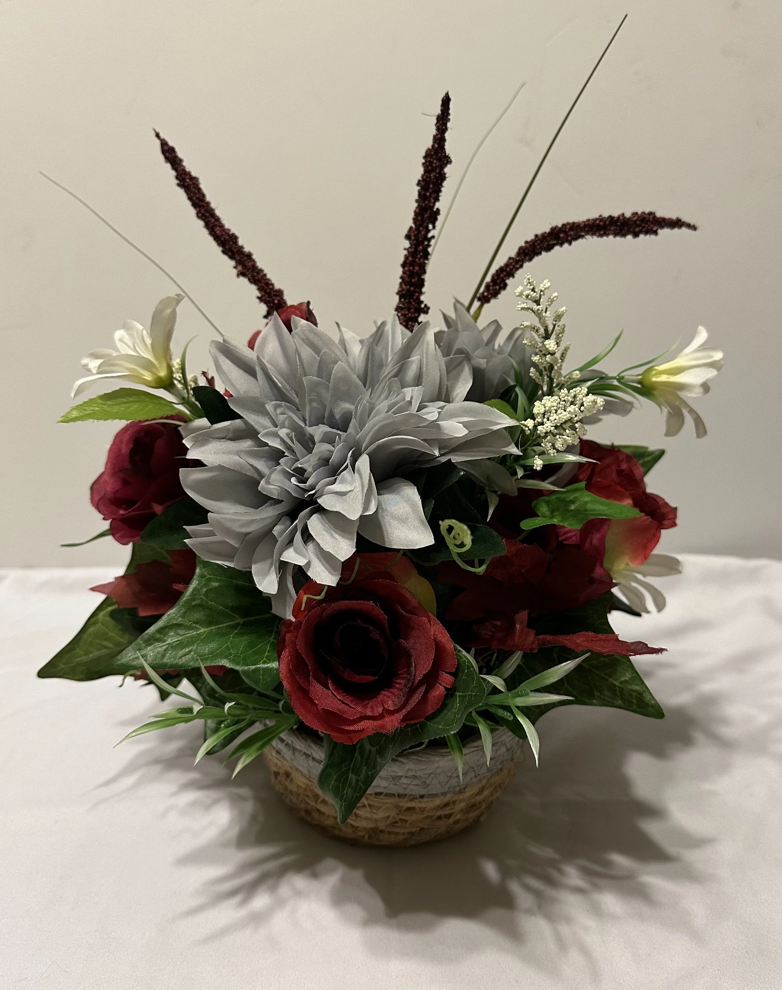Small floral arrangement with red roses, white lilies, a large gray flower, greenery, and decorative grasses in a woven basket