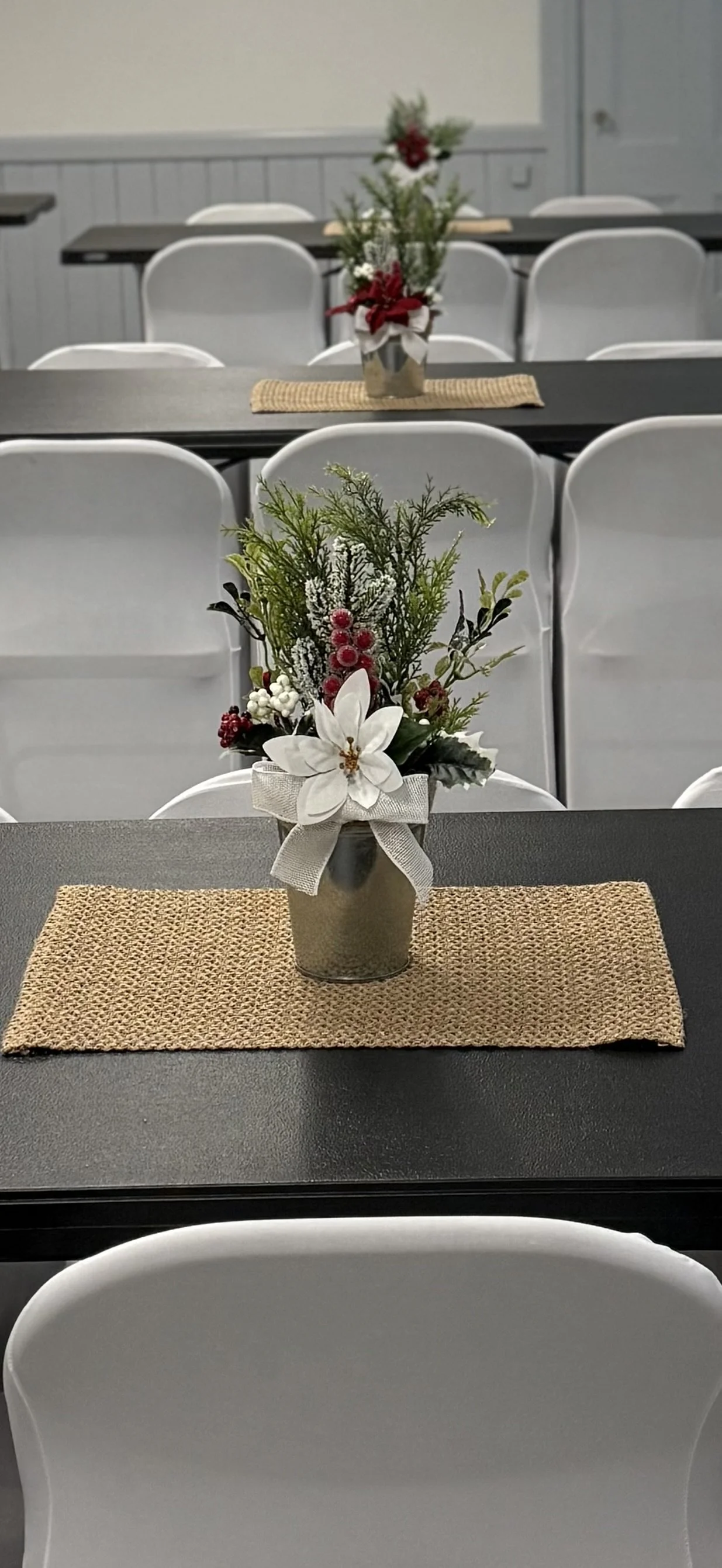 A table decorated for a festive occasion with a small flower arrangement featuring poinsettia, holly, pine, and red berries in a metallic vase with a beige ribbon.