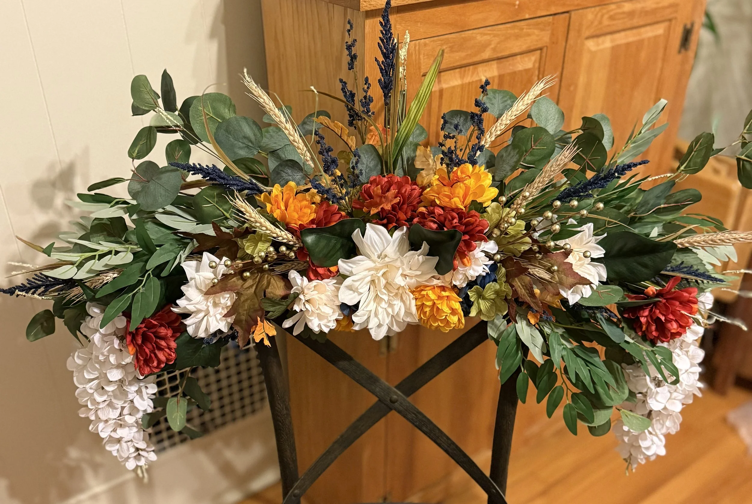Holiday floral swag with white, orange, red, and yellow flowers, including hydrangeas and dahlias, accented by green leaves, purple sprigs, and wheat stalks.