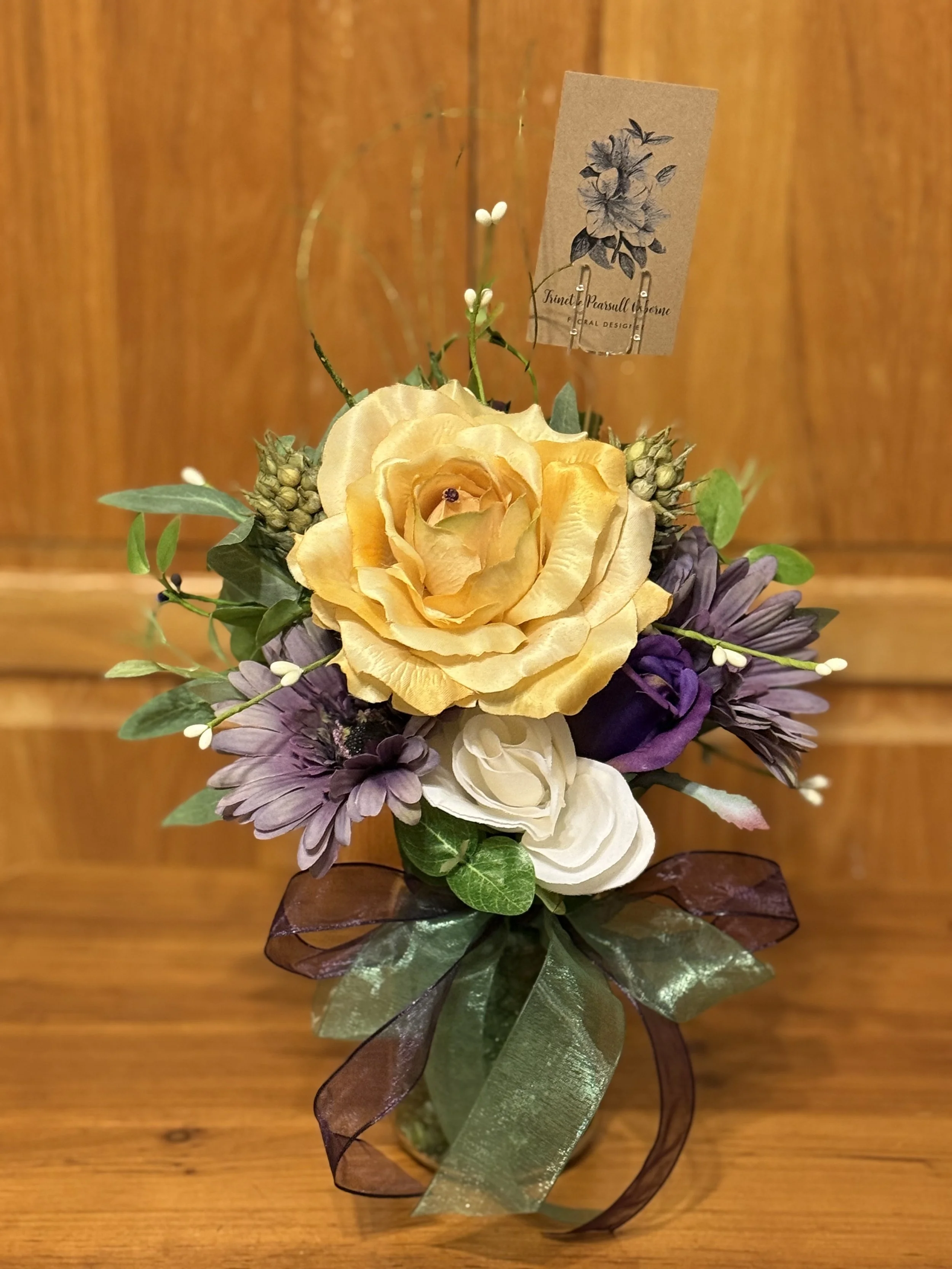 A small floral arrangement in a glass vase with a yellow rose, white roses, purple flowers, and greenery, tied with a brown ribbon, on a wooden surface.