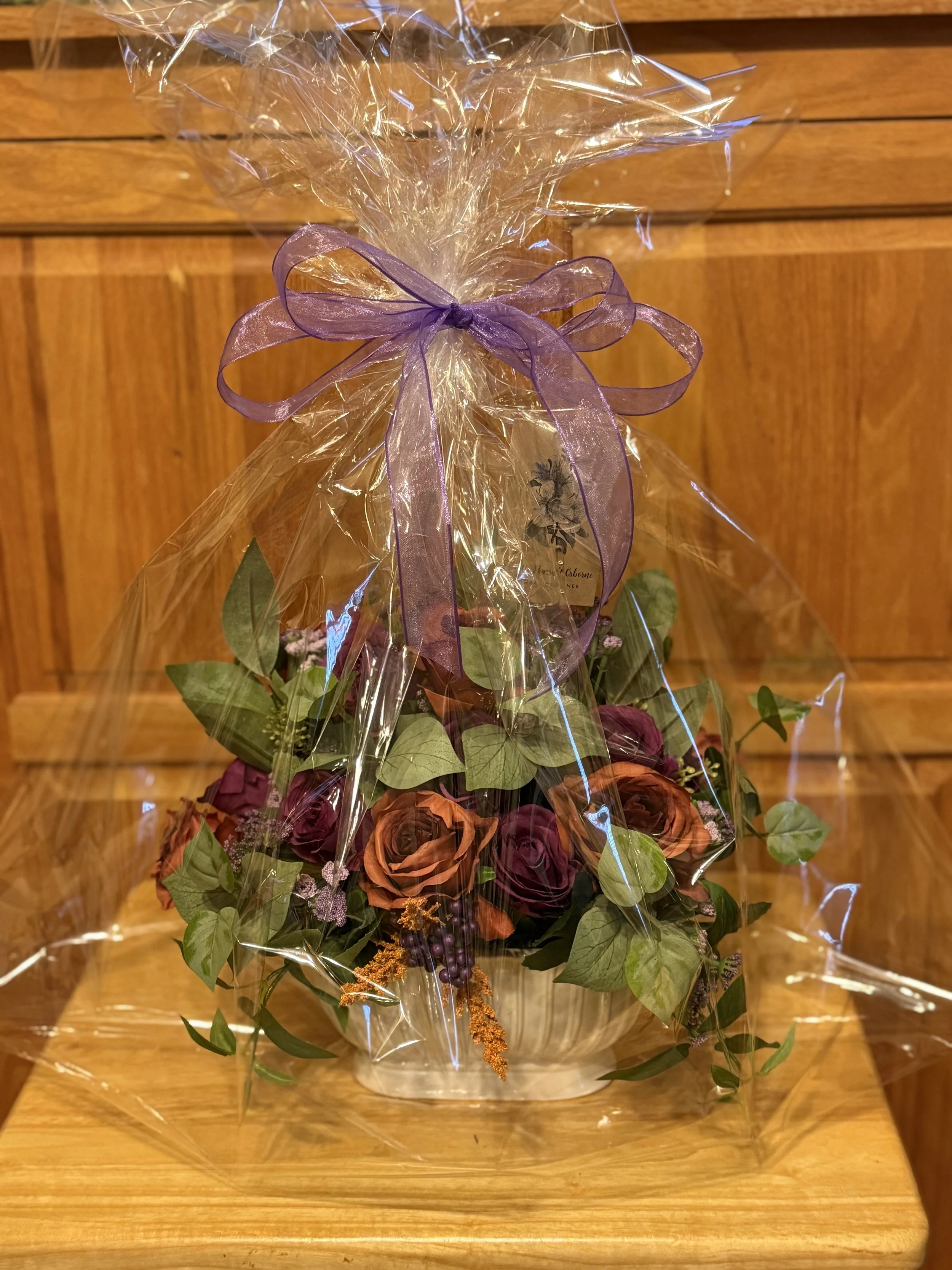 Bouquet of dark purple, and burnt orange roses with green leaves, wrapped in clear cellophane with a purple ribbon.