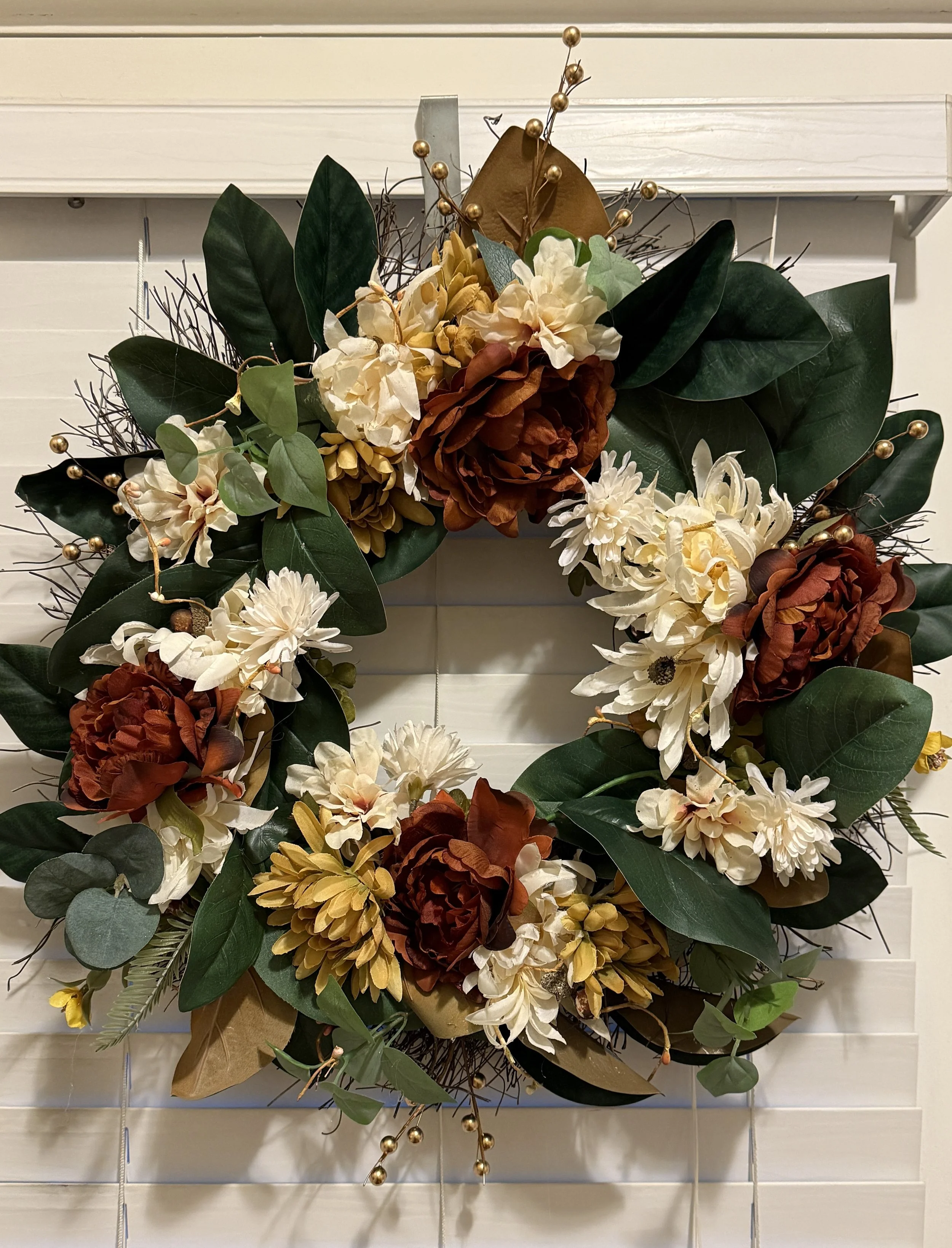 Decorative floral wreath with cream, orange, yellow flowers, large green leaves, and gold accents, hanging on a white wooden wall.