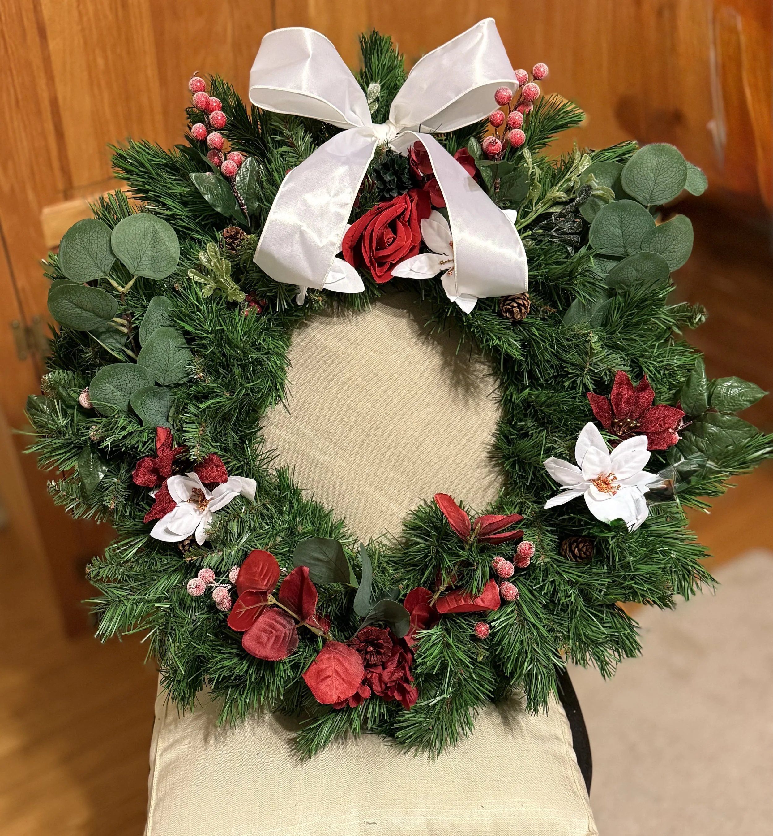 Decorative Christmas wreath with red and white flowers, green pine branches, berries, pinecones, and a white bow.