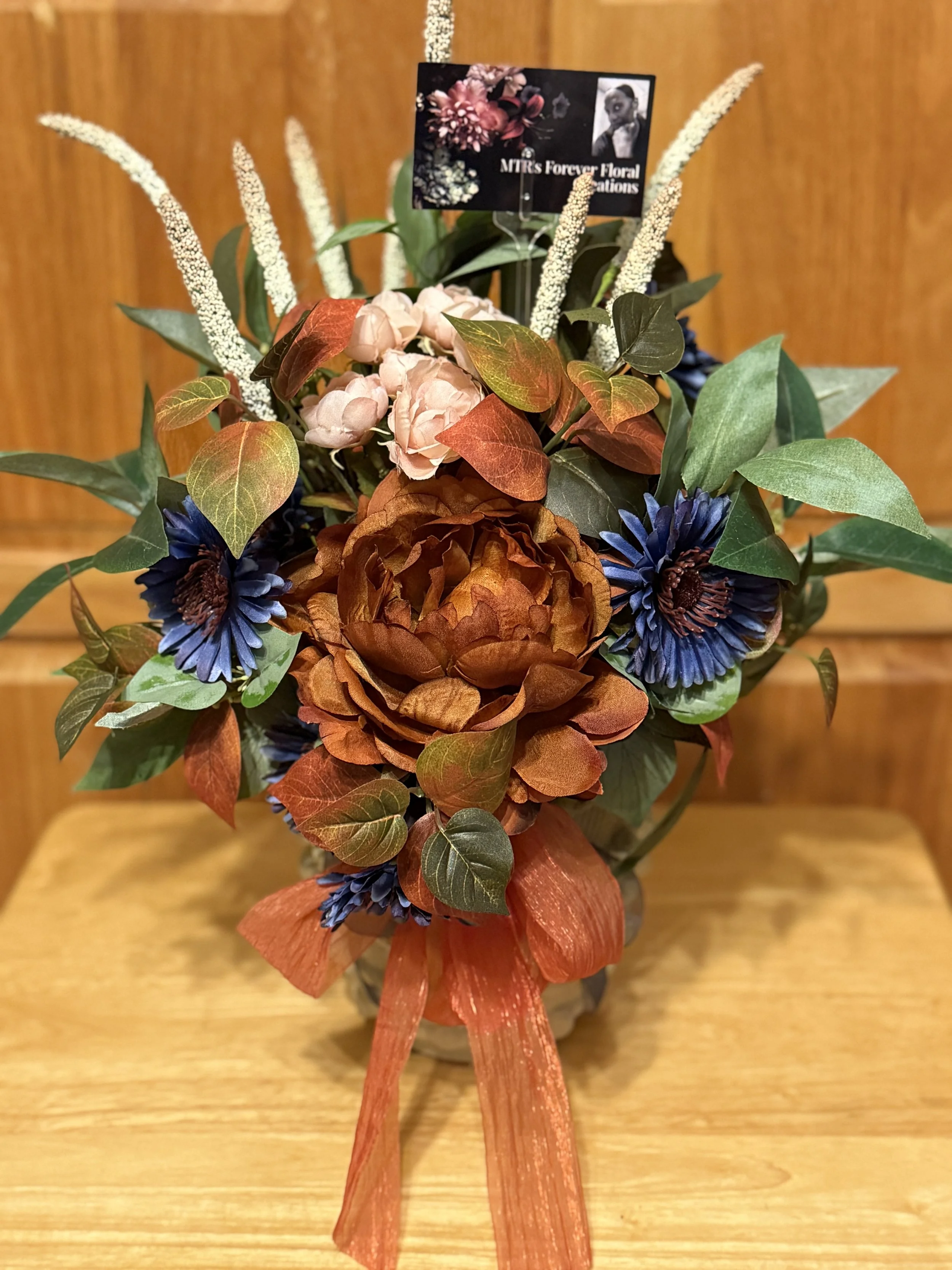 A floral arrangement with pink hydrangeas, blue daisies, brown-orange peonies, green leaves, white streamers, and a black tag that reads 'MTR's Forever Floral Creations,' placed on a wooden table and background.