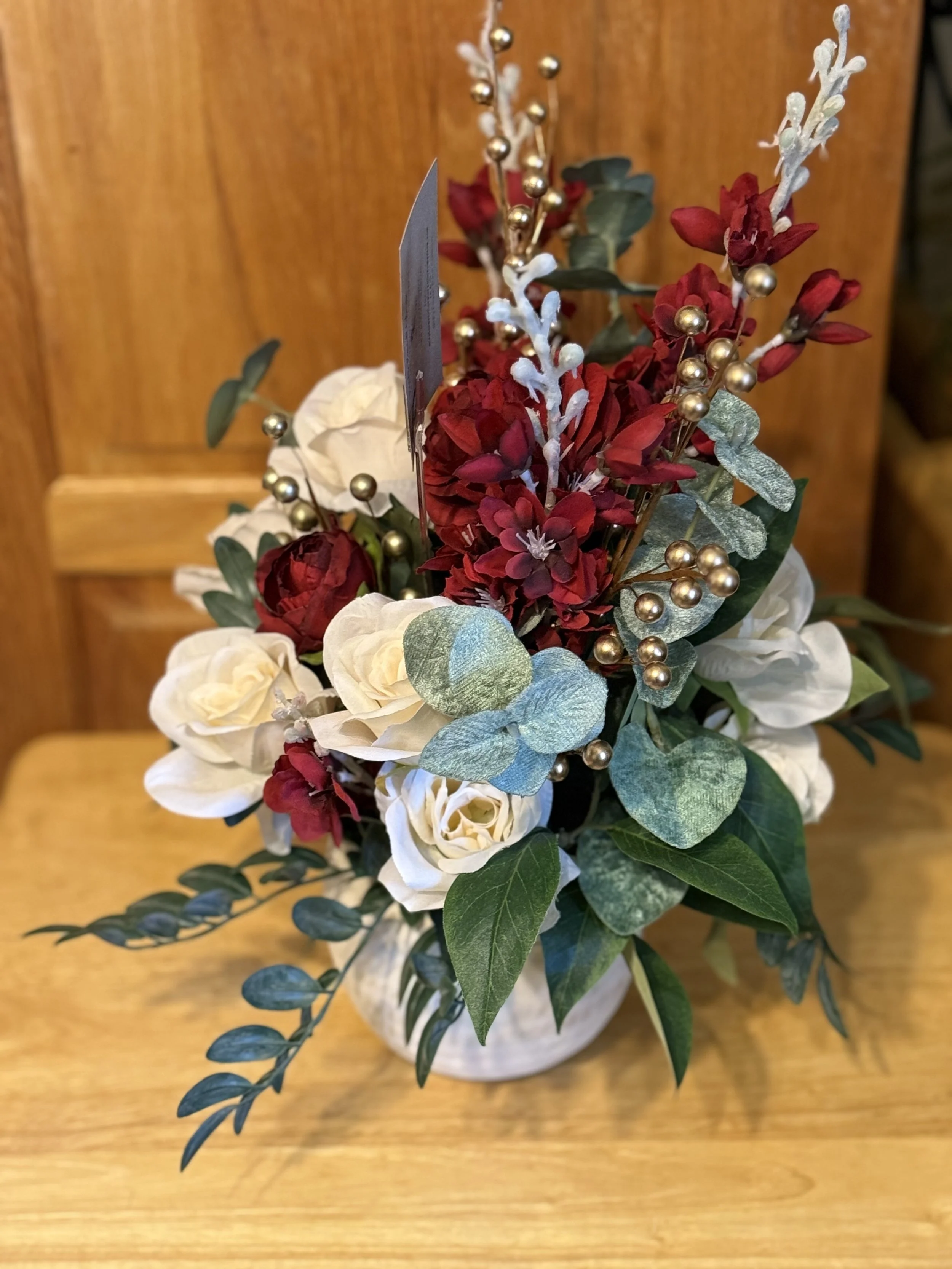Festive floral arrangement with red and white flowers, green leaves, and gold decorative accents in a white vase on a wooden table.