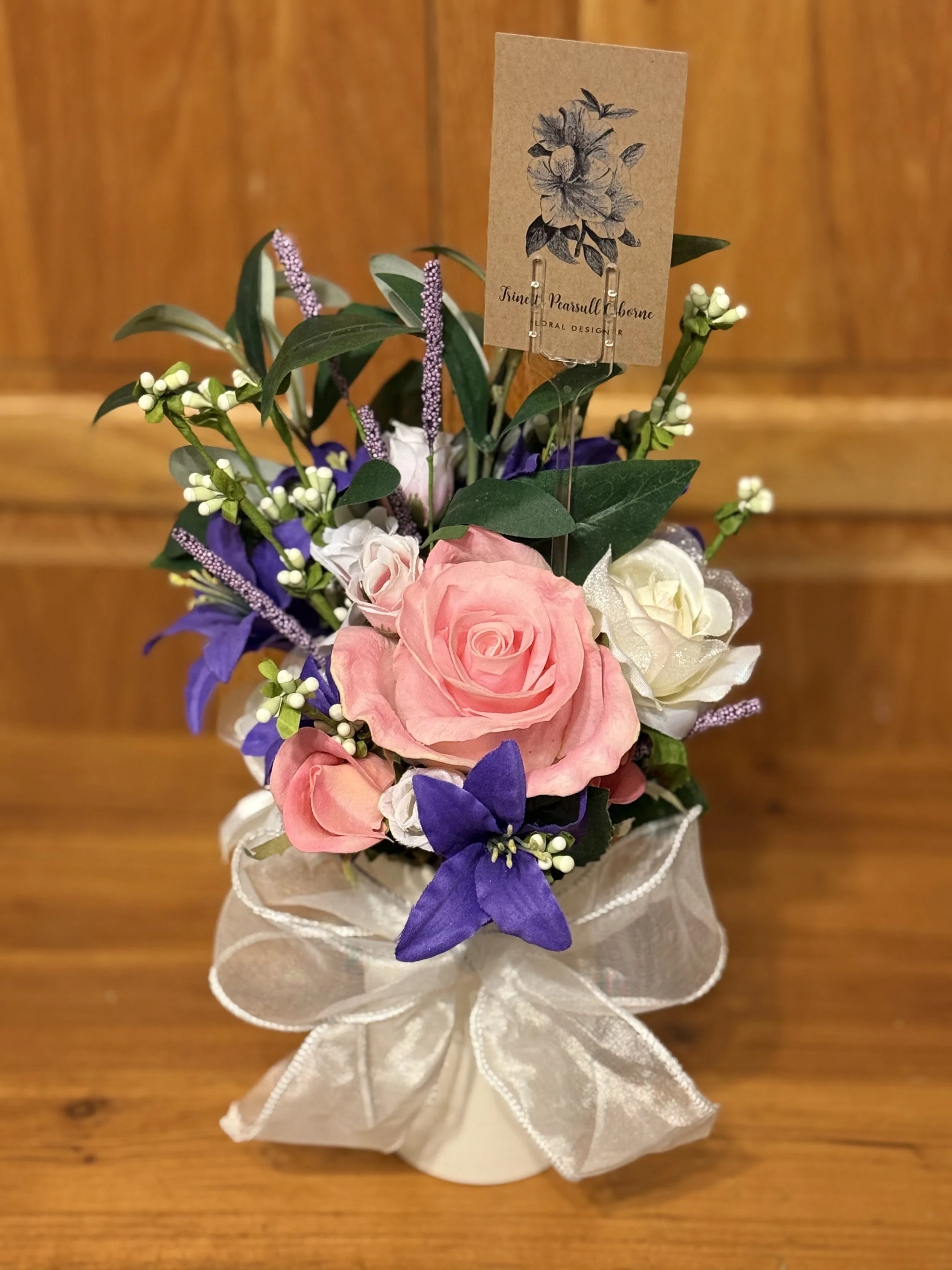 Arrangement of pink, white, and purple flowers in a white pot, wrapped with a sheer ribbon, with a cardboard tag featuring a floral sketch.