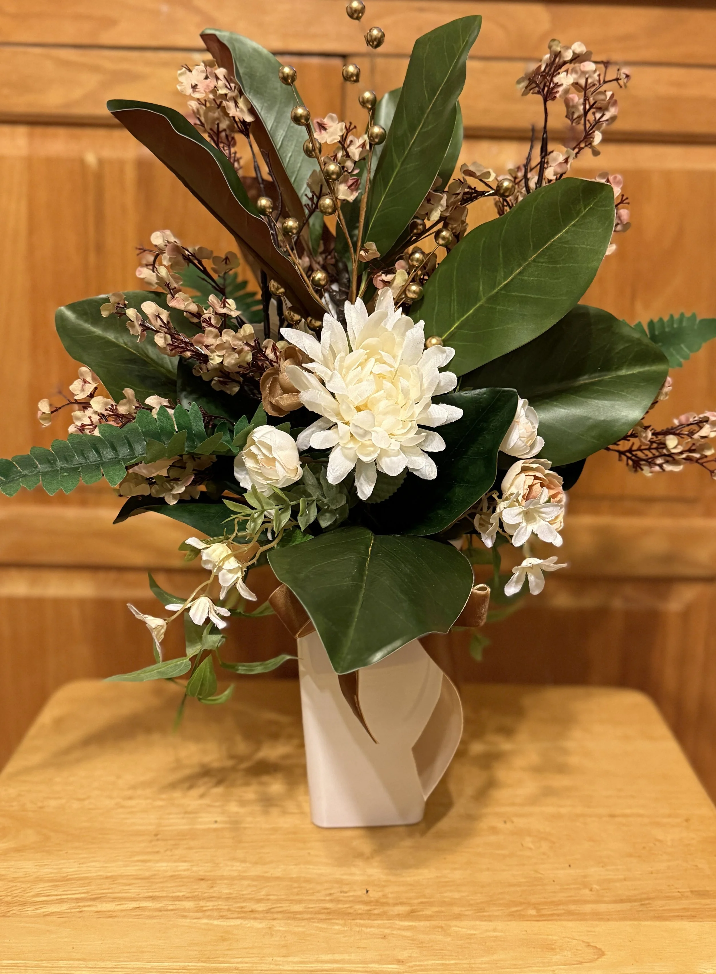 An elegant fall floral arrangement in a white vase containing greenery, white flowers, and small silk beige-colored baby breathes.