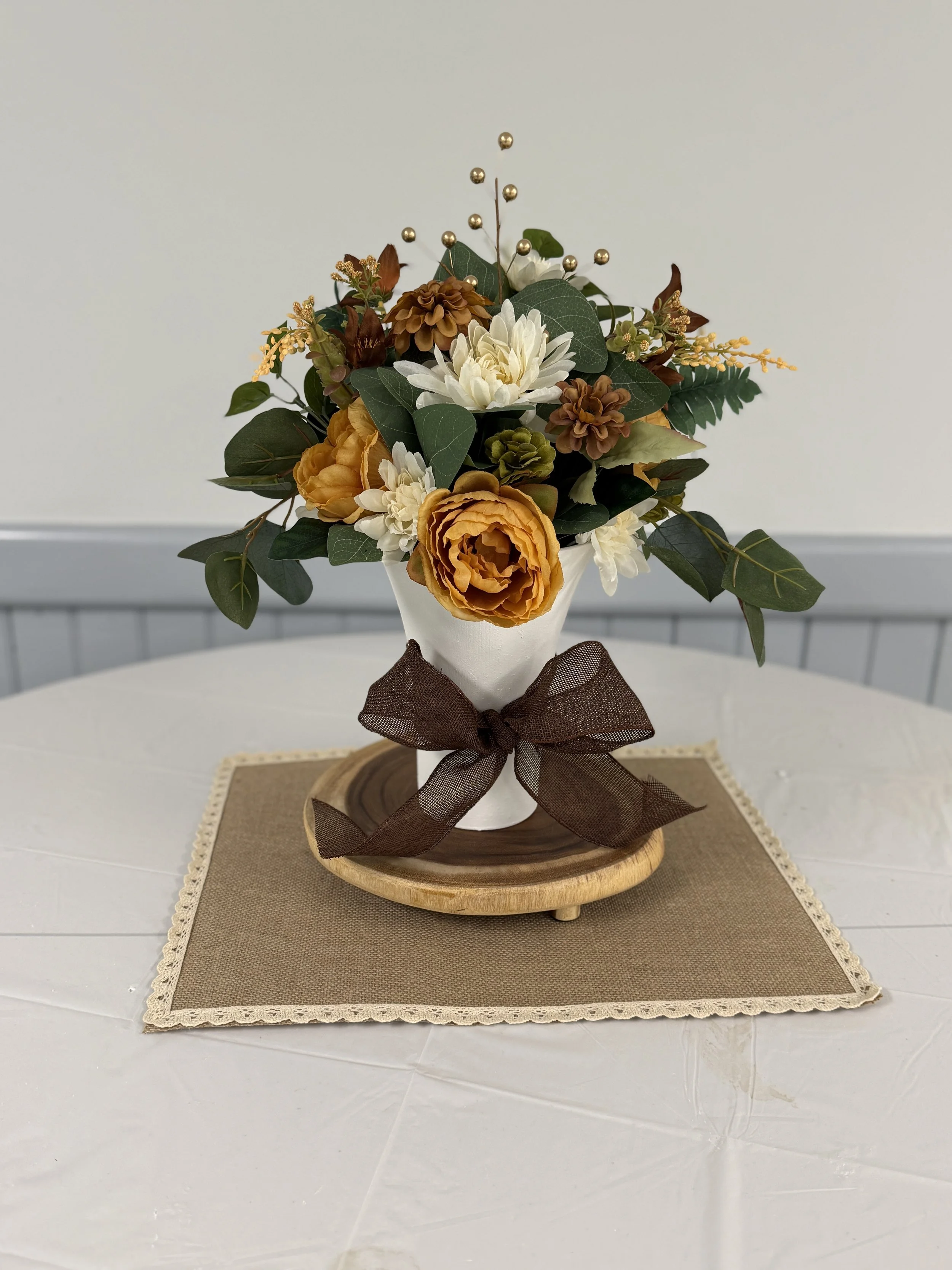 Autumn floral arrangement with mustard, white, and brown flowers in a white vase, decorated with a brown bow, placed on a wooden tray on a beige cloth with lace edges, on a white table.
