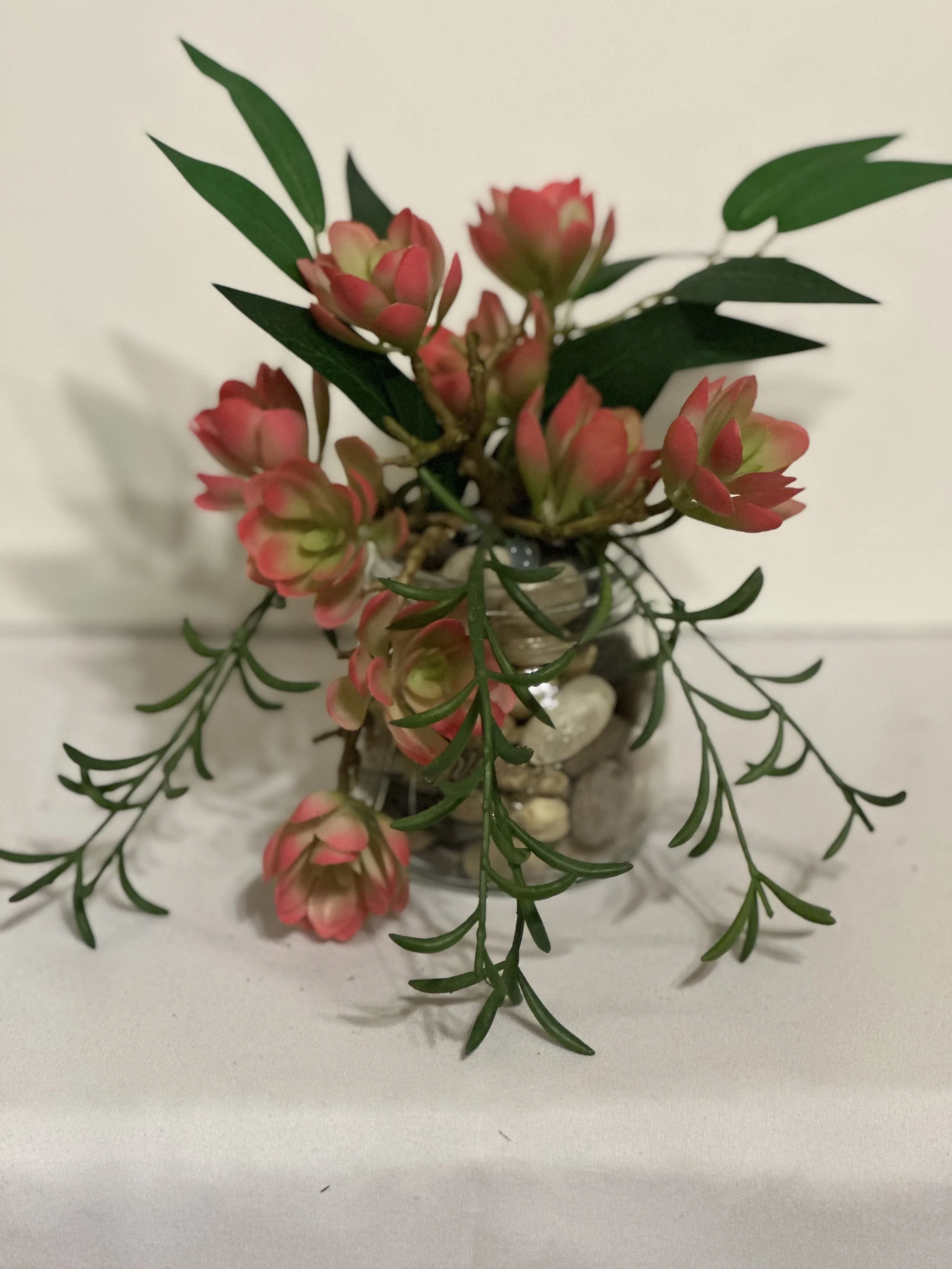 A small glass vase with pink and green succulent plants and green leaves, decorated with small stones inside, on a white surface against a light background.