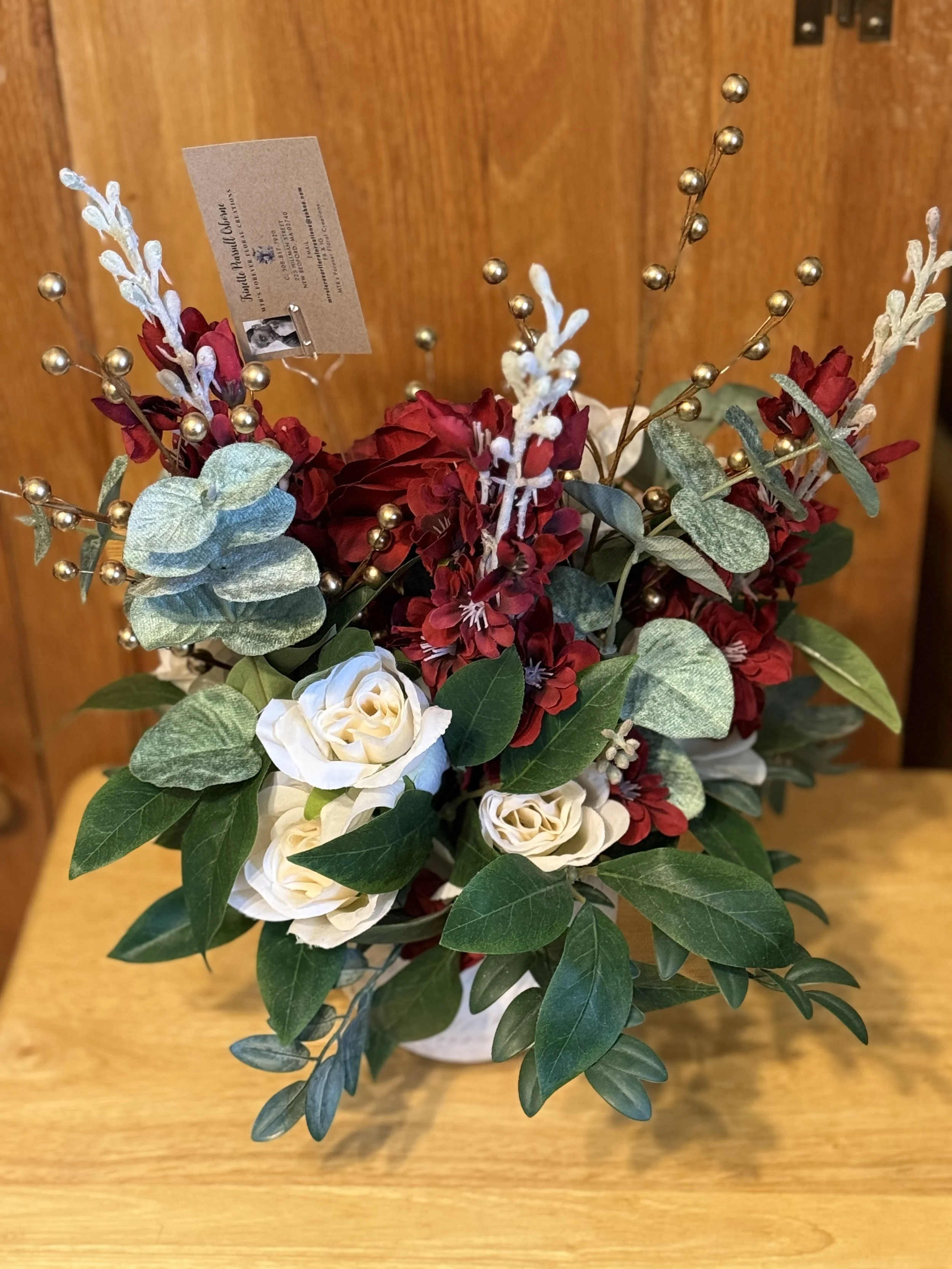 A floral arrangement with white roses, red flowers, green leaves, and decorative gold berry branches, placed on a wooden surface against a wooden background.