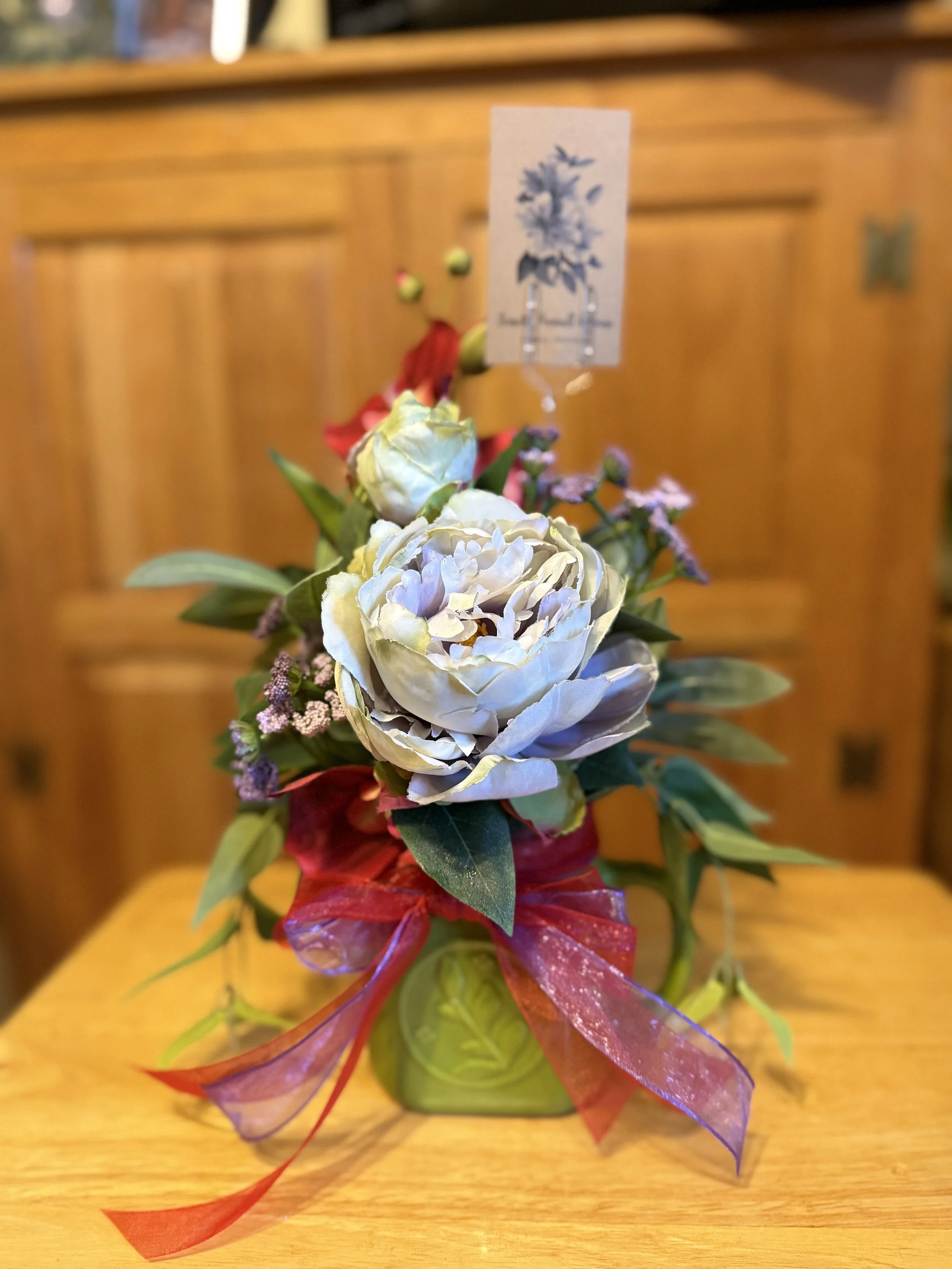 A small floral arrangement in a green vase with a red ribbon bow, featuring white peonies, purple statice, and green foliage. A white tag with blue floral design is attached to the arrangement.