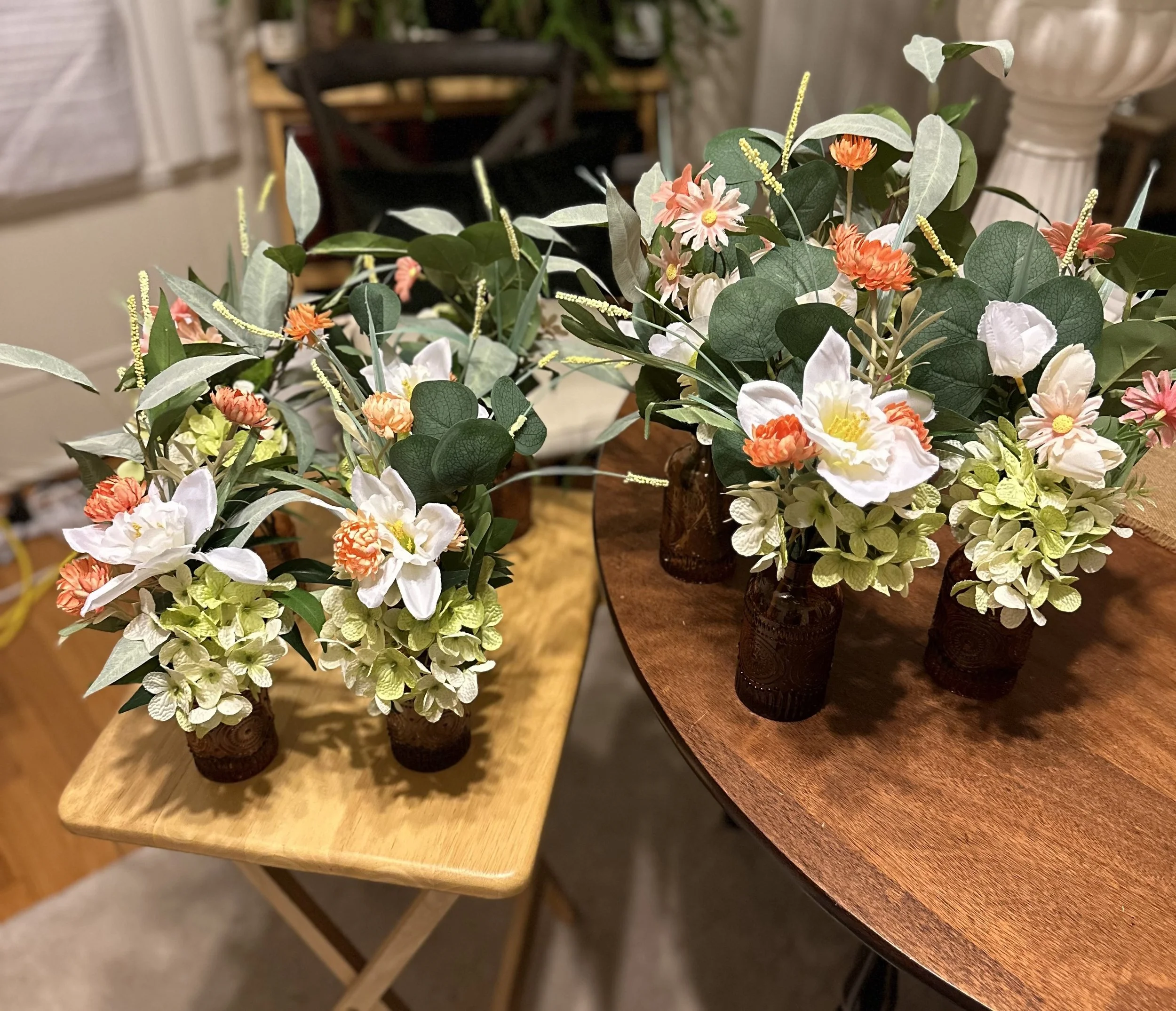Two floral arrangements in small brown glass vases sitting on wooden tables, one with a round edge and the other with a rectangular edge, featuring white, peach, and pink flowers with green leaves and foliage.