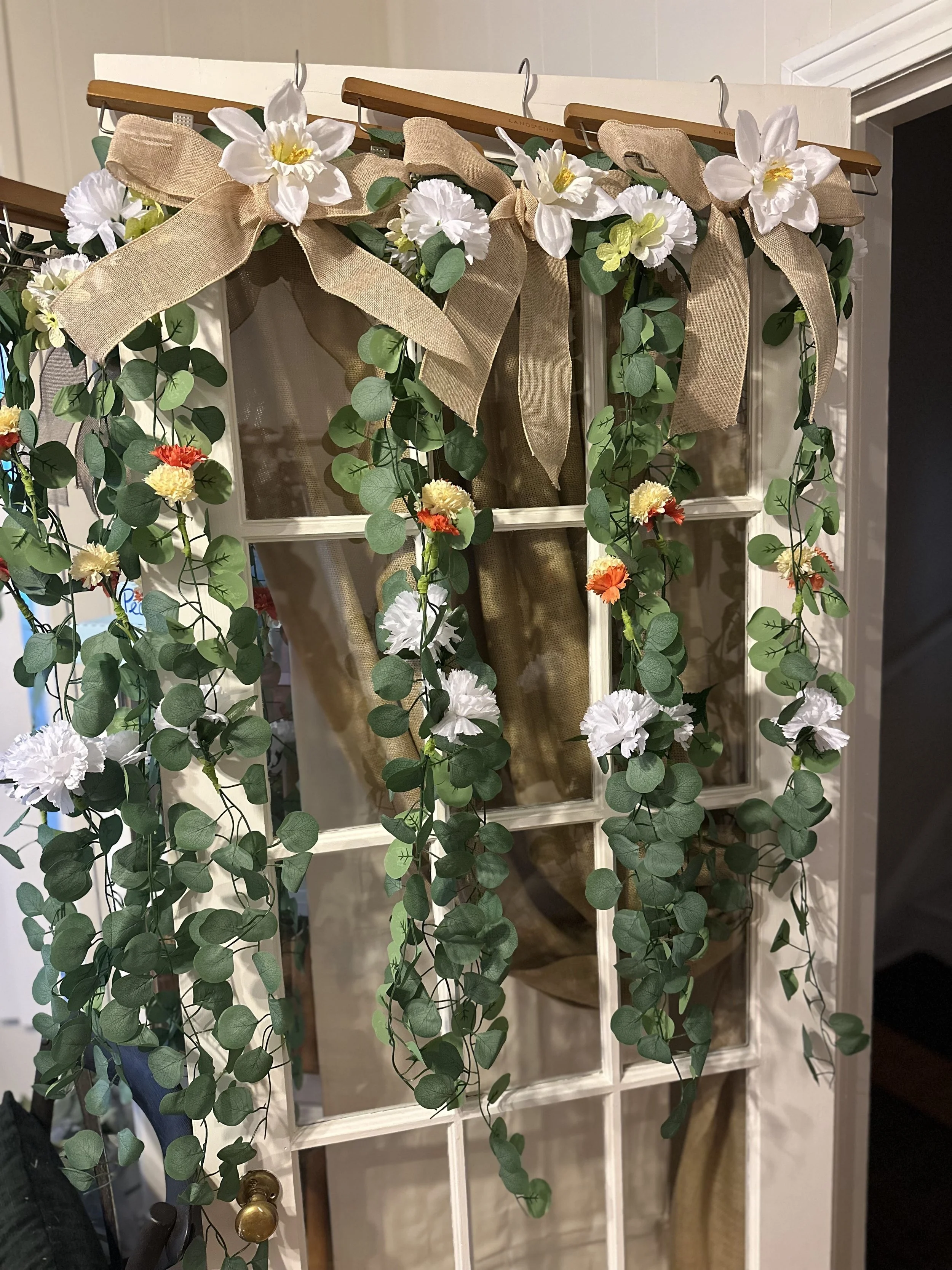 Decorative door hanging with artificial flowers, greenery, and large beige fabric bows.